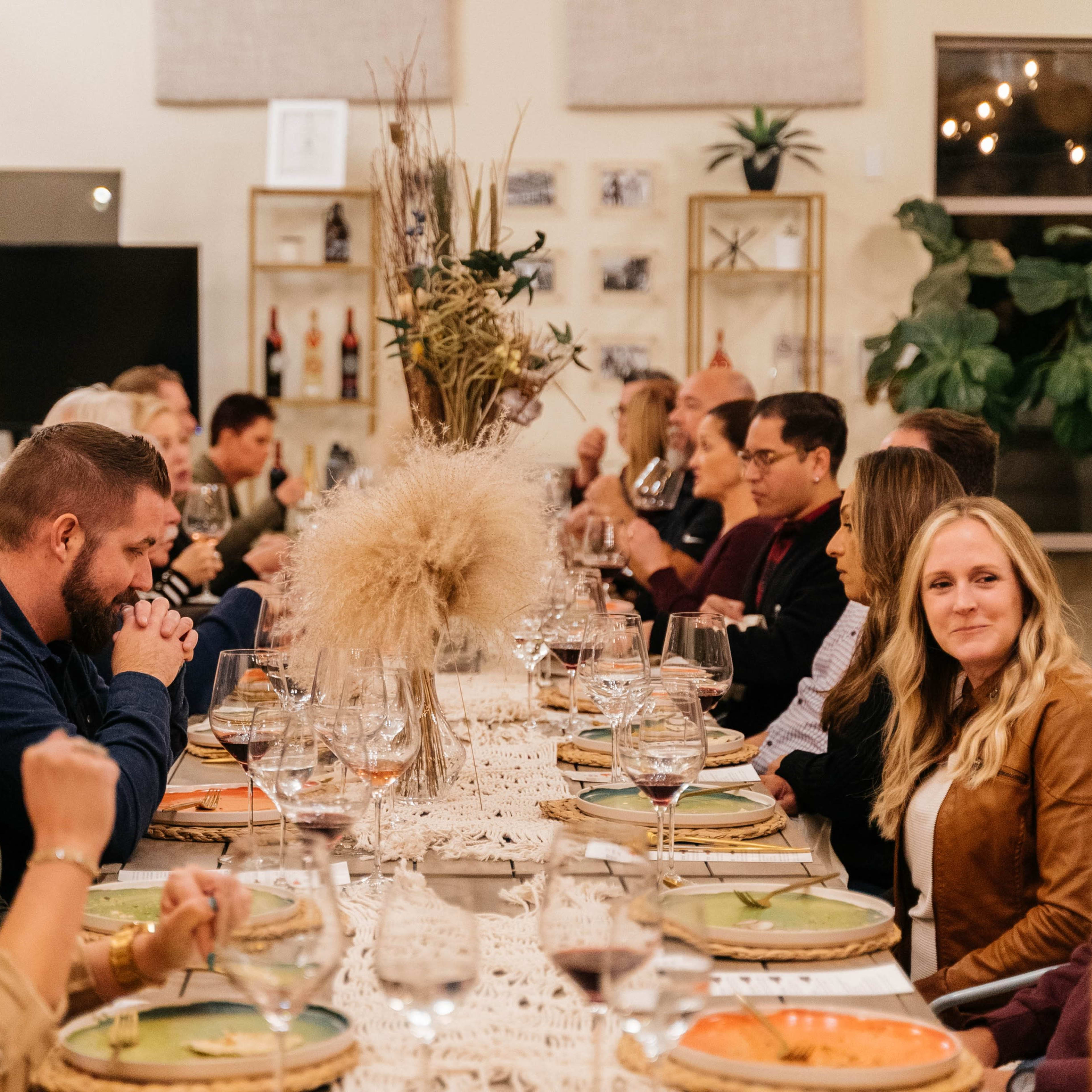 A long dining table is set with plates and glasses, and a group of guests are seated on either side, engaged in conversation during a meal.