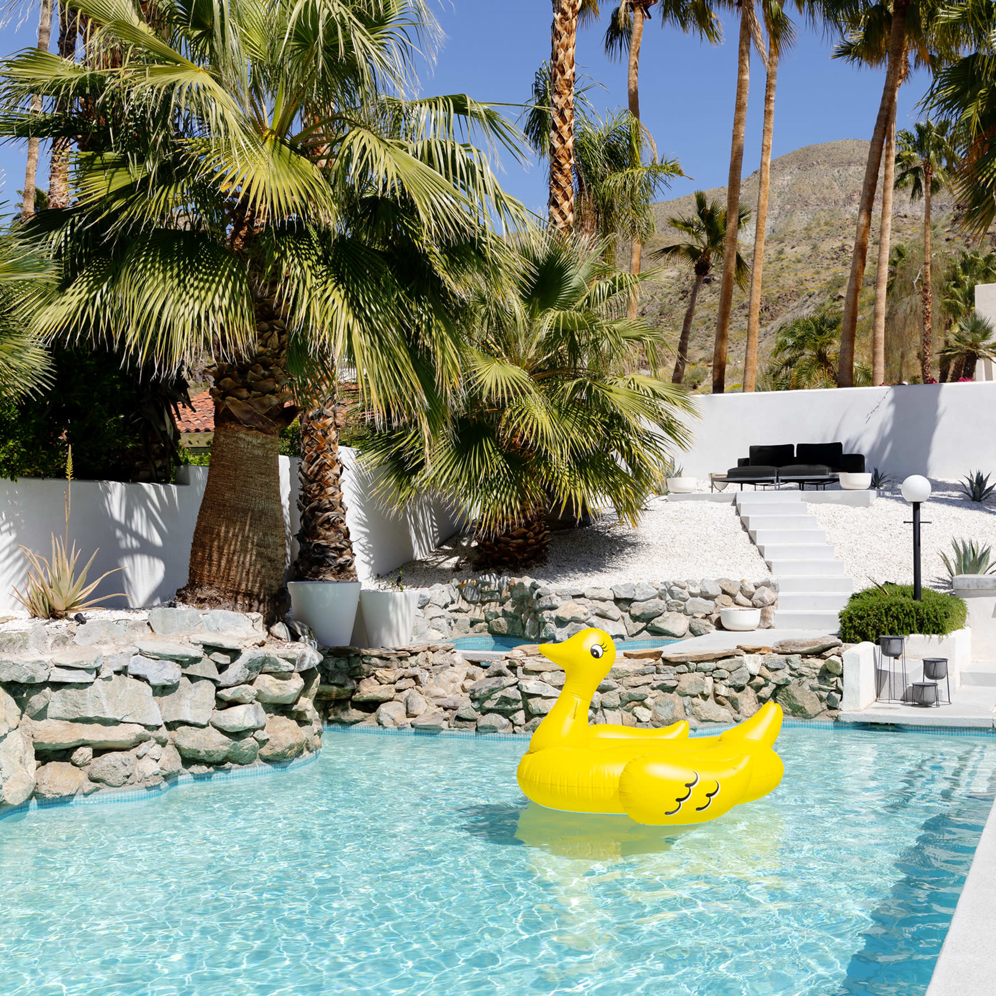 A large yellow duck-shaped float is positioned in a clear swimming pool surrounded by palm trees and stone landscaping.