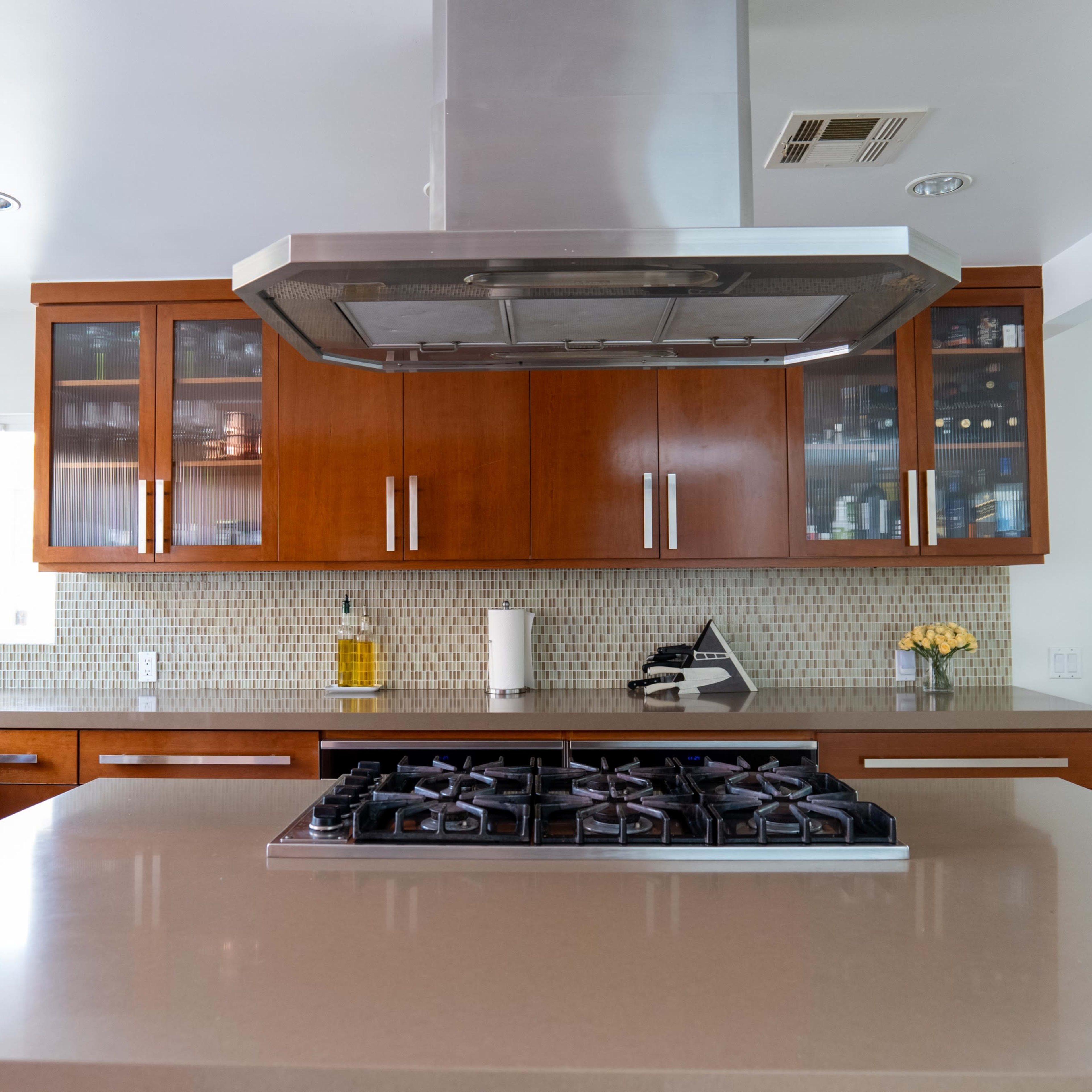 A modern kitchen with wooden cabinetry, a central island featuring a gas stove, and a silver range hood above it.