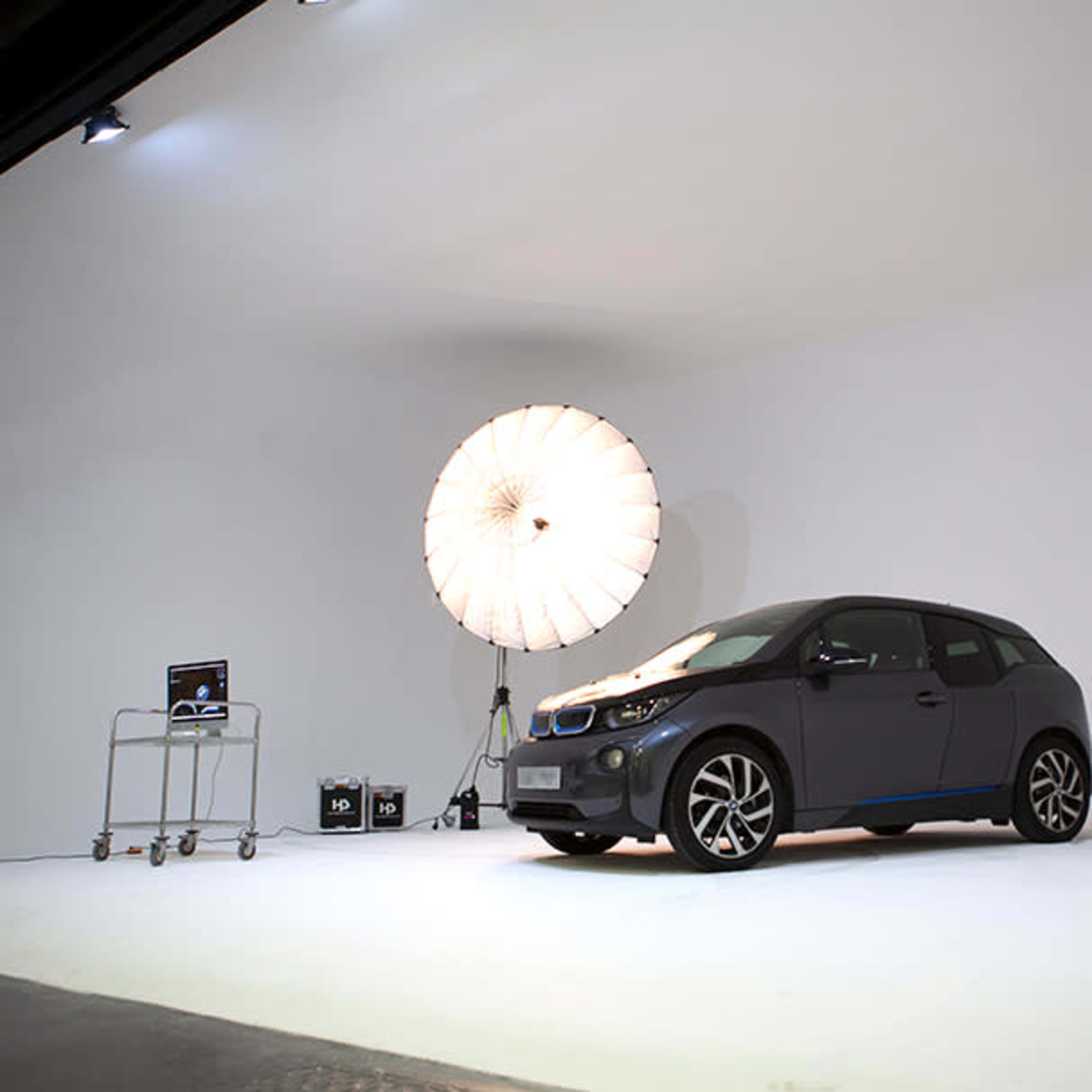 A dark gray electric car is positioned on a white backdrop in a studio, with a large softbox light illuminating the scene.