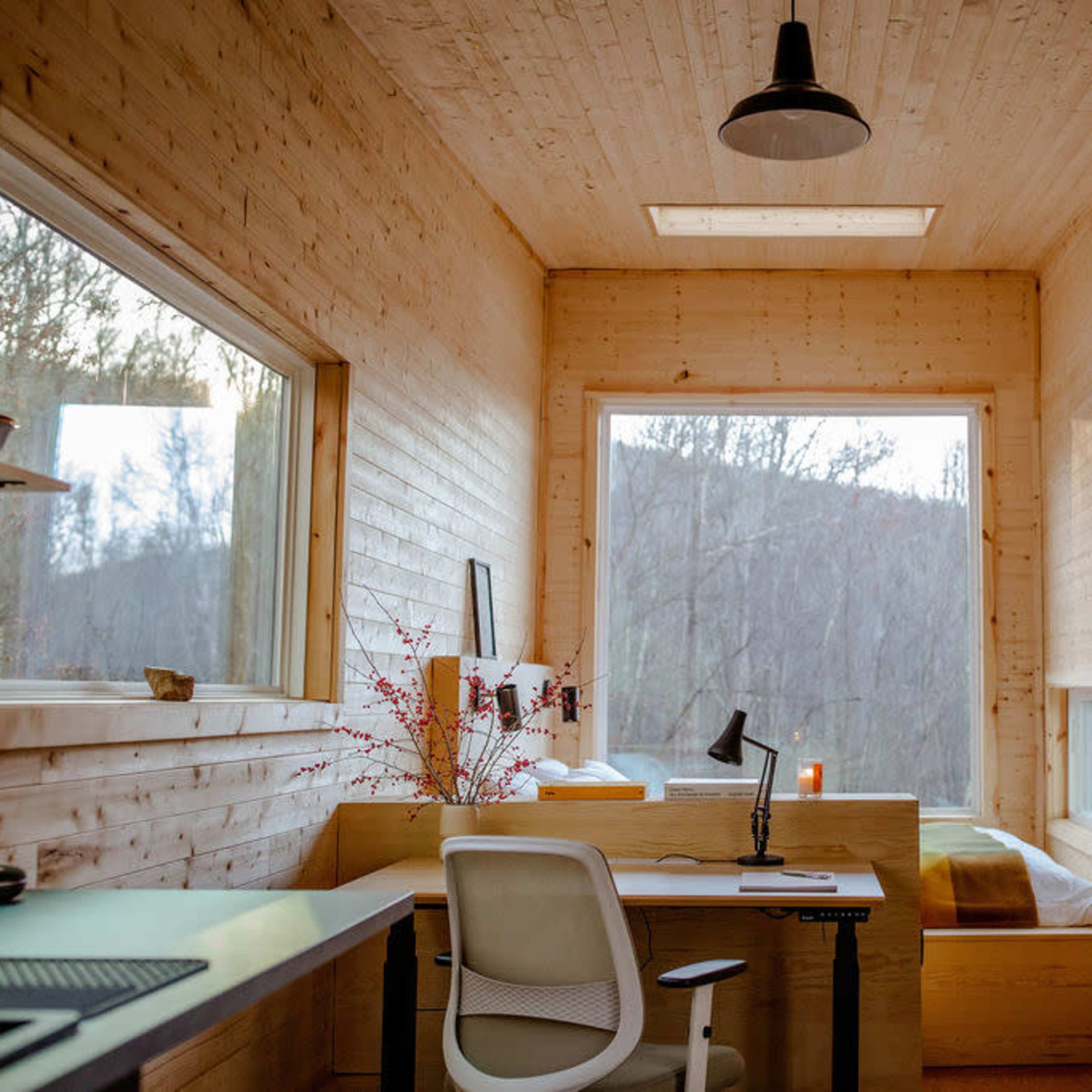 A minimalist wooden interior of a small room with a desk, chair, and a bed next to large windows that overlook a forested area.