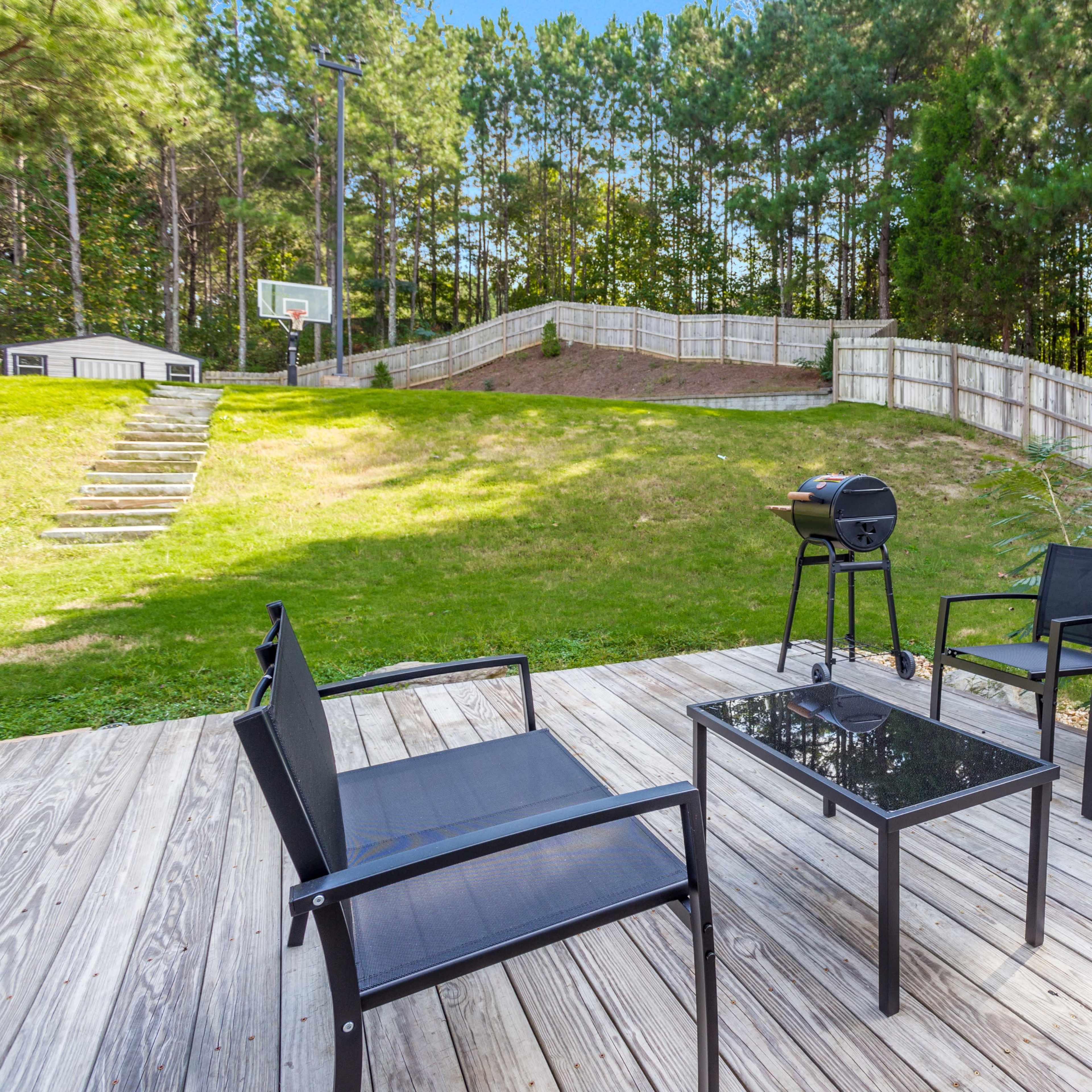 A wooden deck features a black patio table and chairs, overlooking a grassy yard with a charcoal grill and basketball hoop in the background.