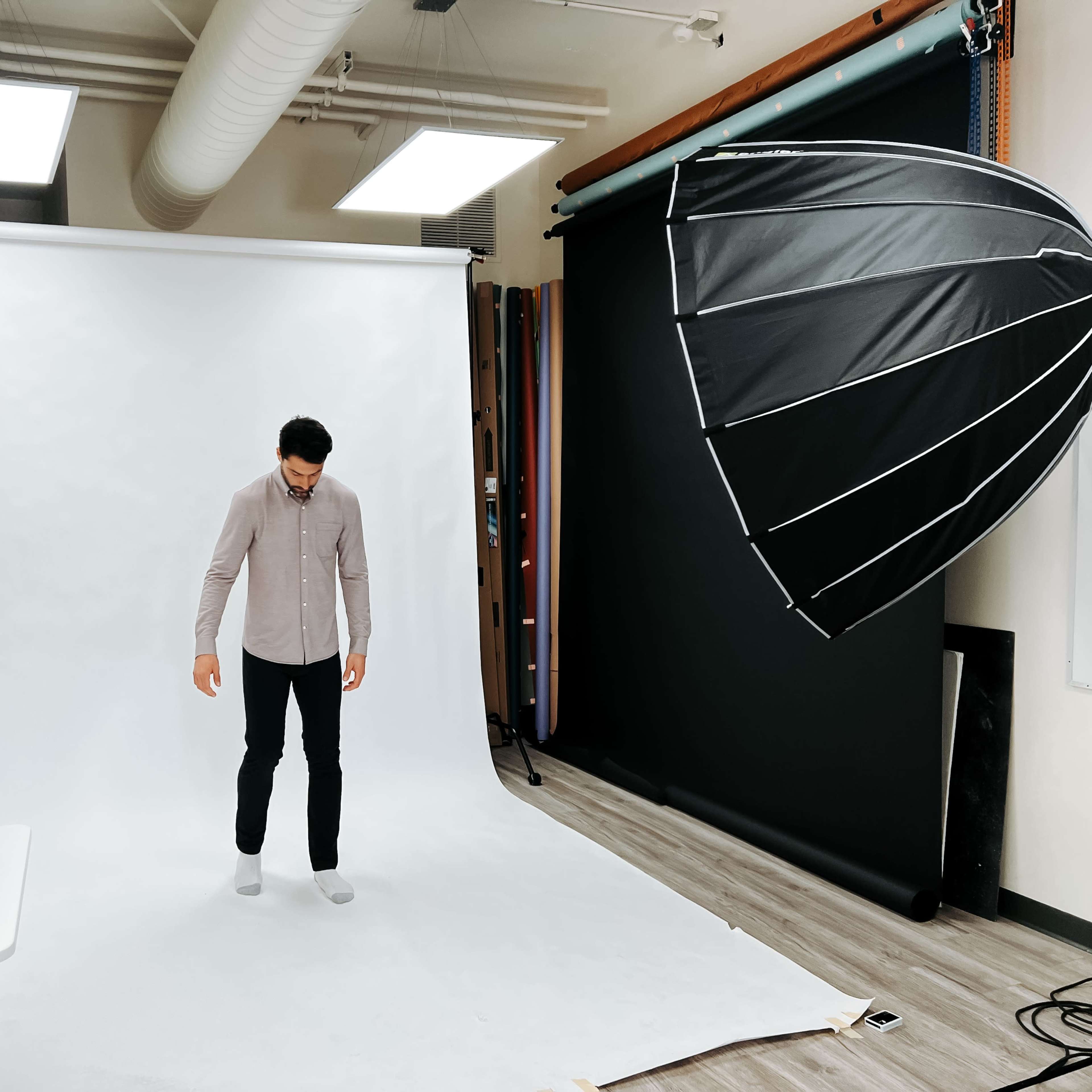 A person stands on a light-colored backdrop in a photography studio, with an umbrella light positioned to the side.