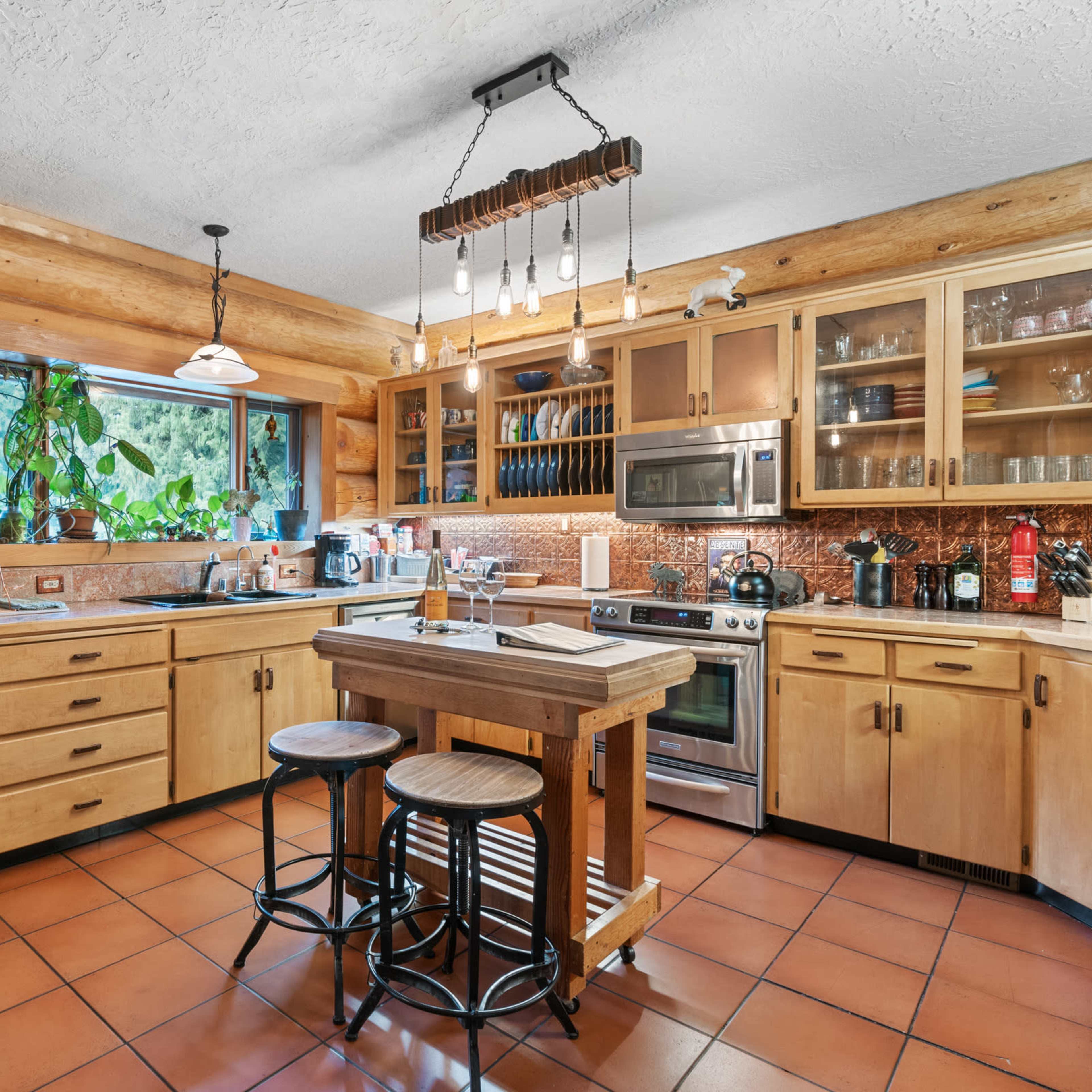 A rustic kitchen features wooden cabinets, a stainless steel refrigerator, a large island in the center, and a tiled floor with ample natural light from a window.