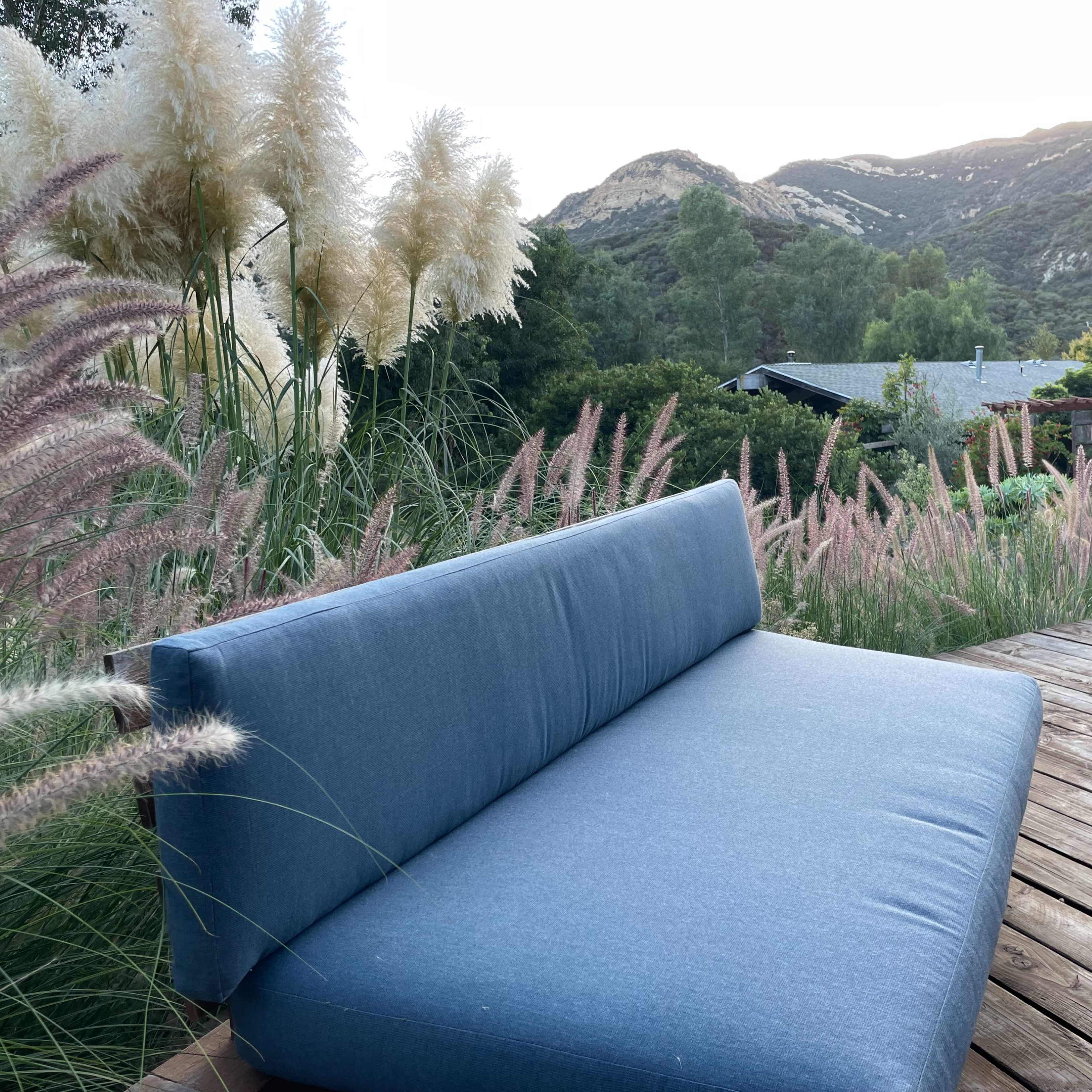 A blue outdoor sofa is positioned on a wooden deck surrounded by tall grasses and mountains in the background.