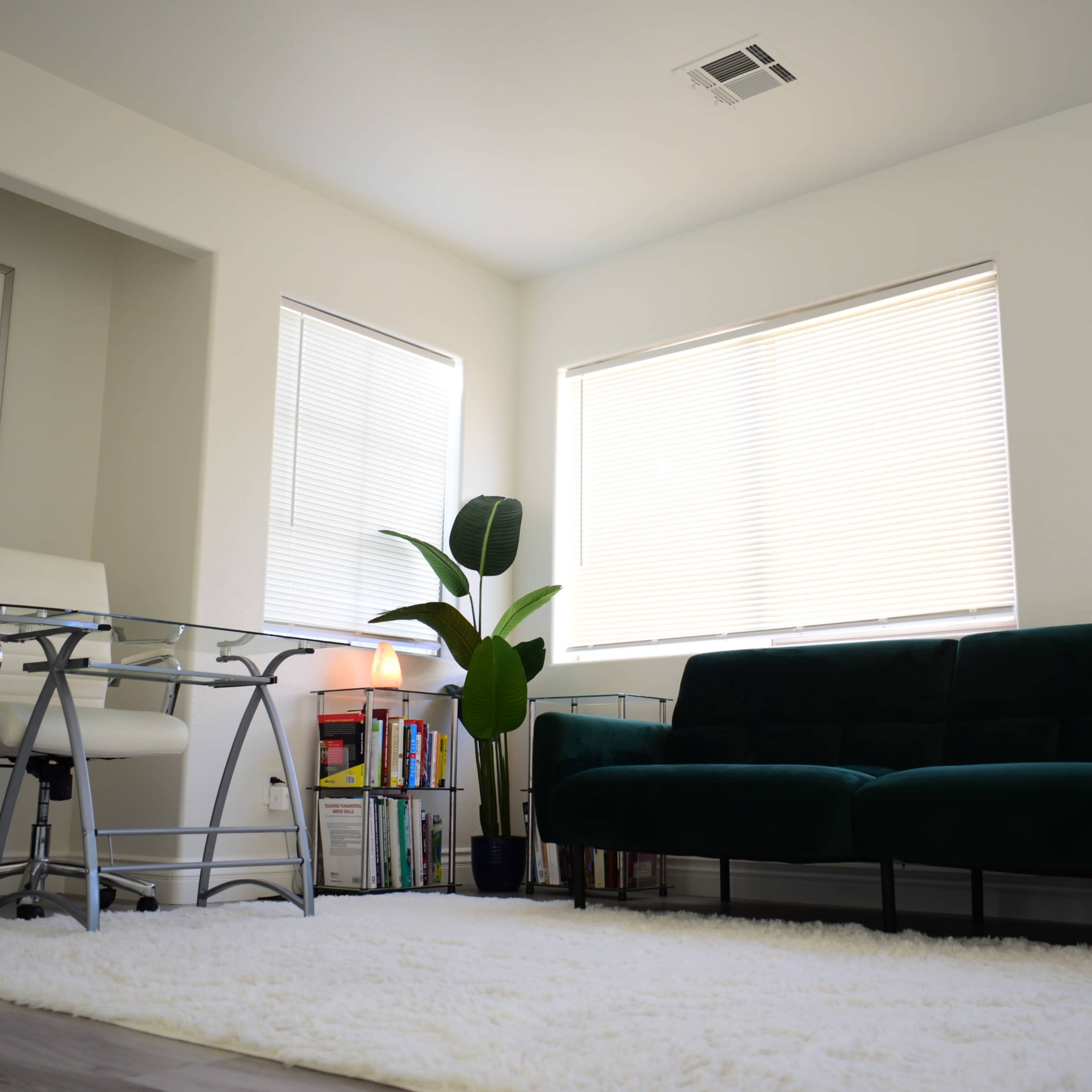 A well-lit room features a green sofa, a glass desk with a chair, and artwork on the walls, with sunlight streaming through the windows.