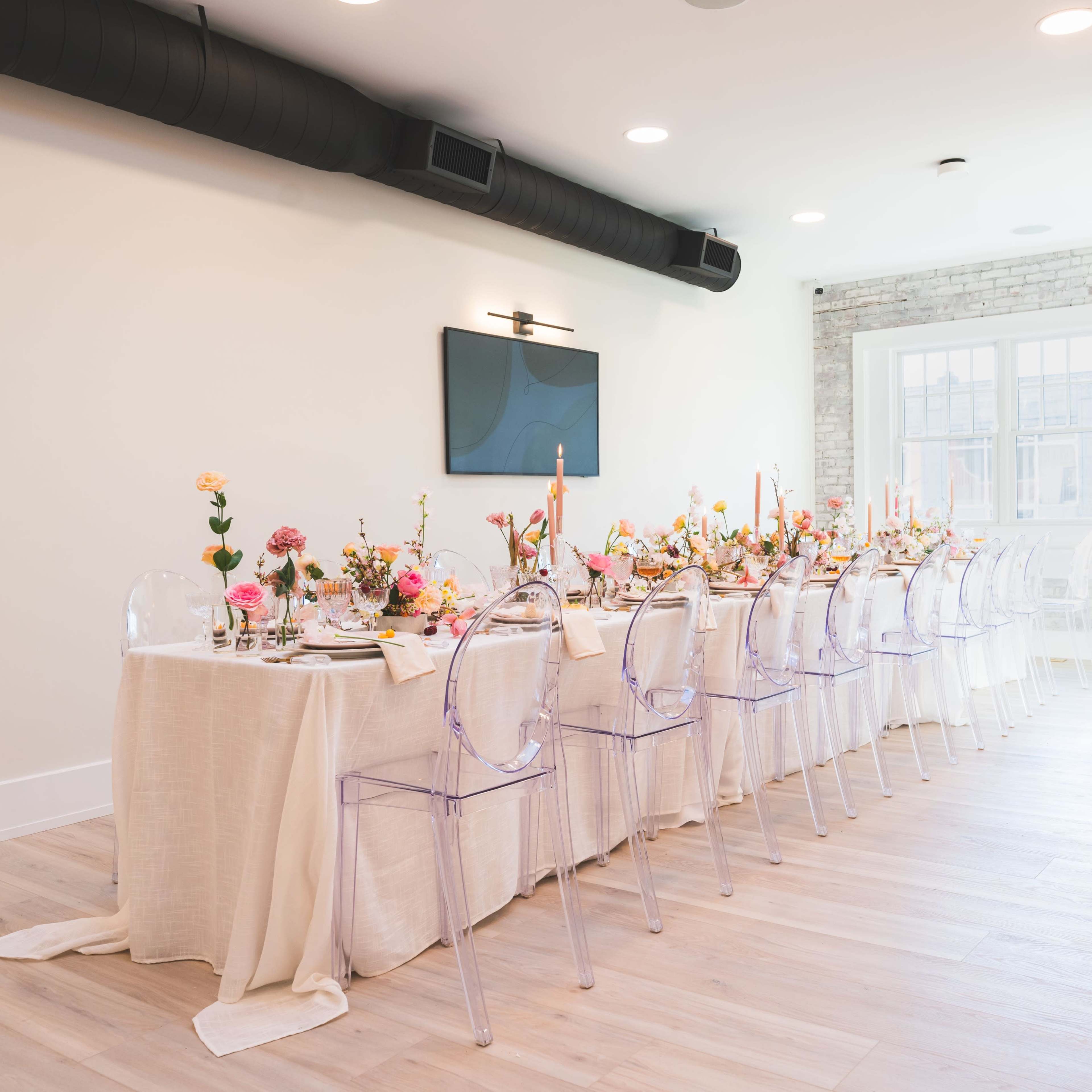 A long dining table decorated with flowers and candles sits under a modern light fixture in a bright room with a large window.