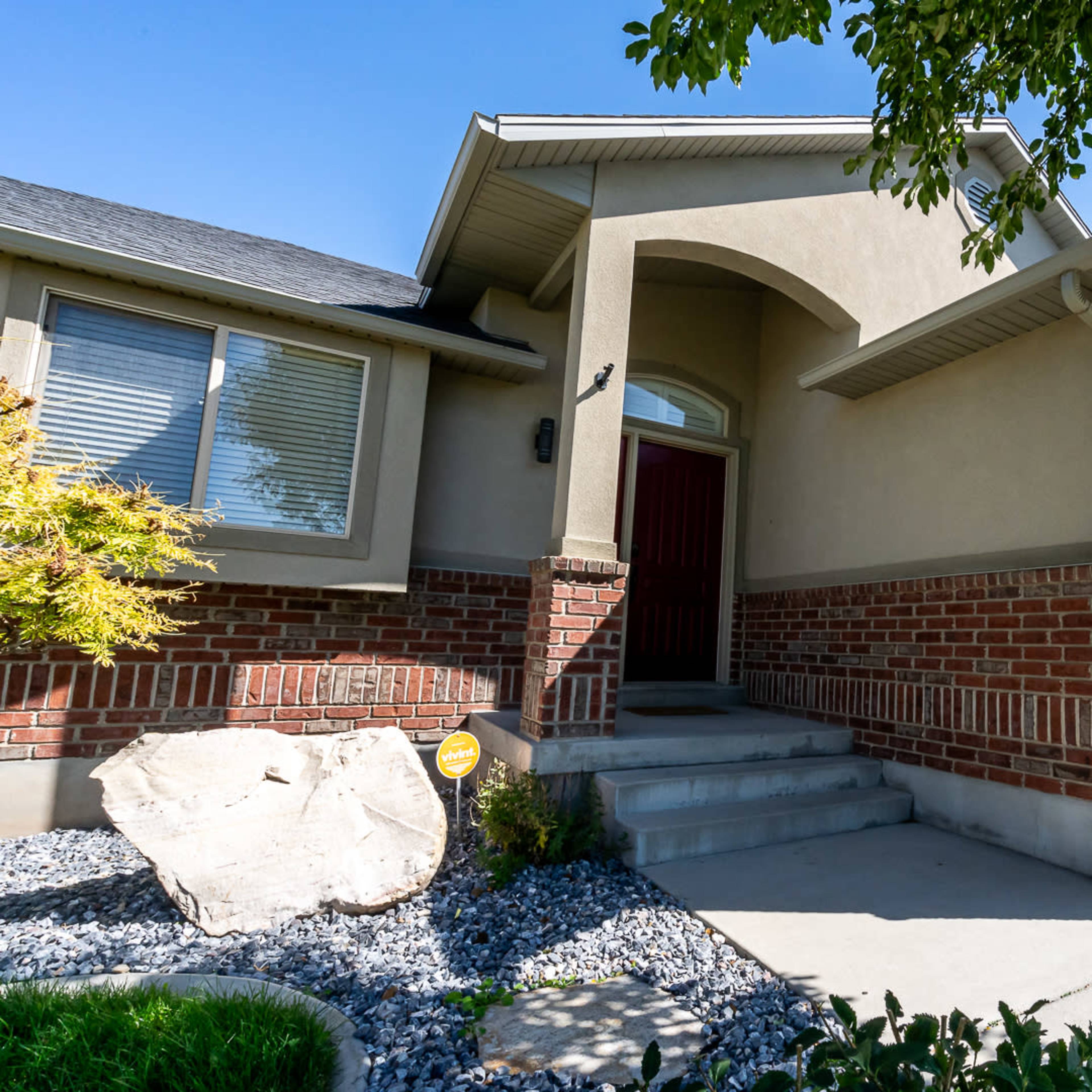 The entrance of a single-story house featuring a brick and stucco exterior, with a landscaped yard and a paved walkway leading to the door.