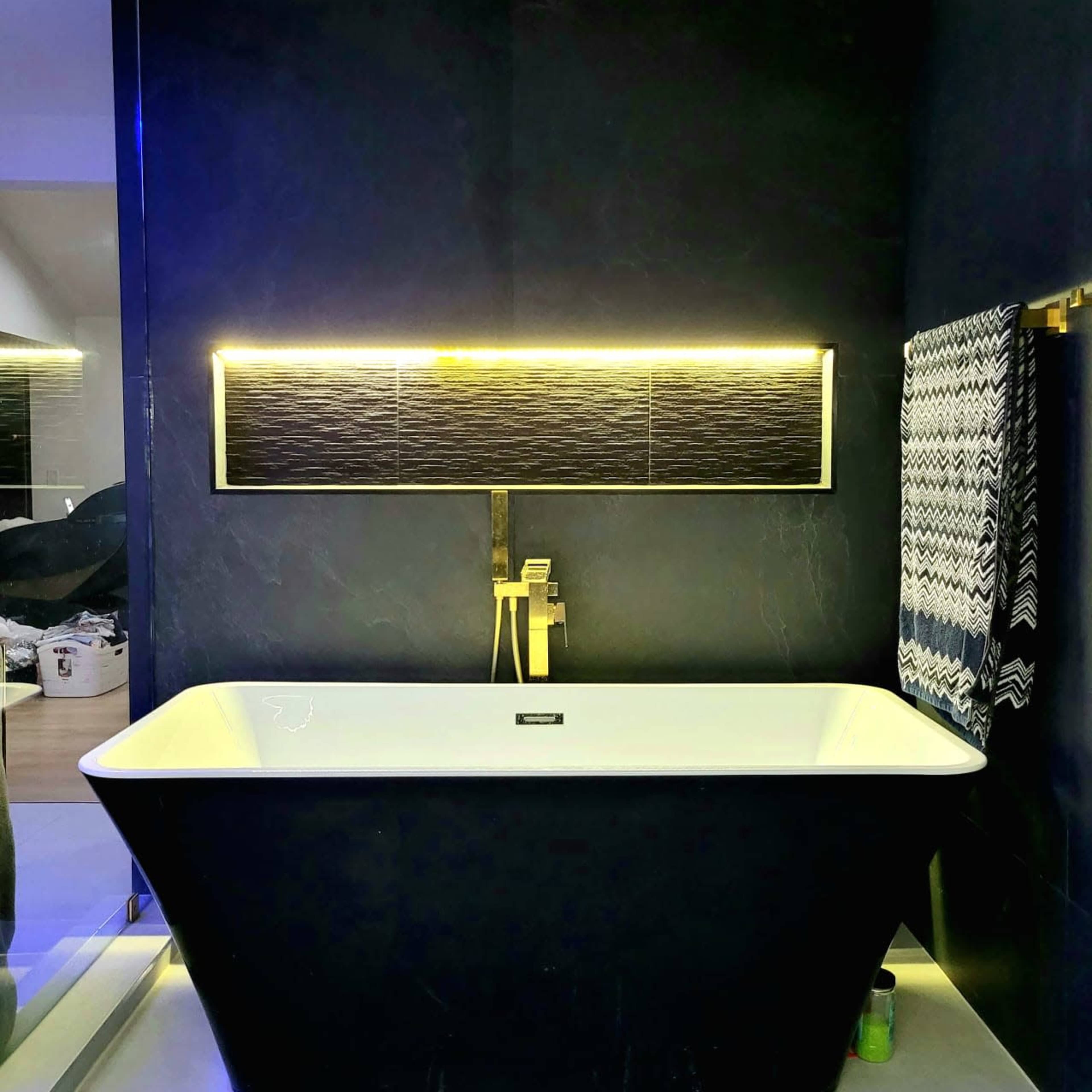 A modern bathroom featuring a sleek black freestanding bathtub, stylish wall-mounted faucet, illuminated wall panel above the tub, and a patterned towel hanging nearby.