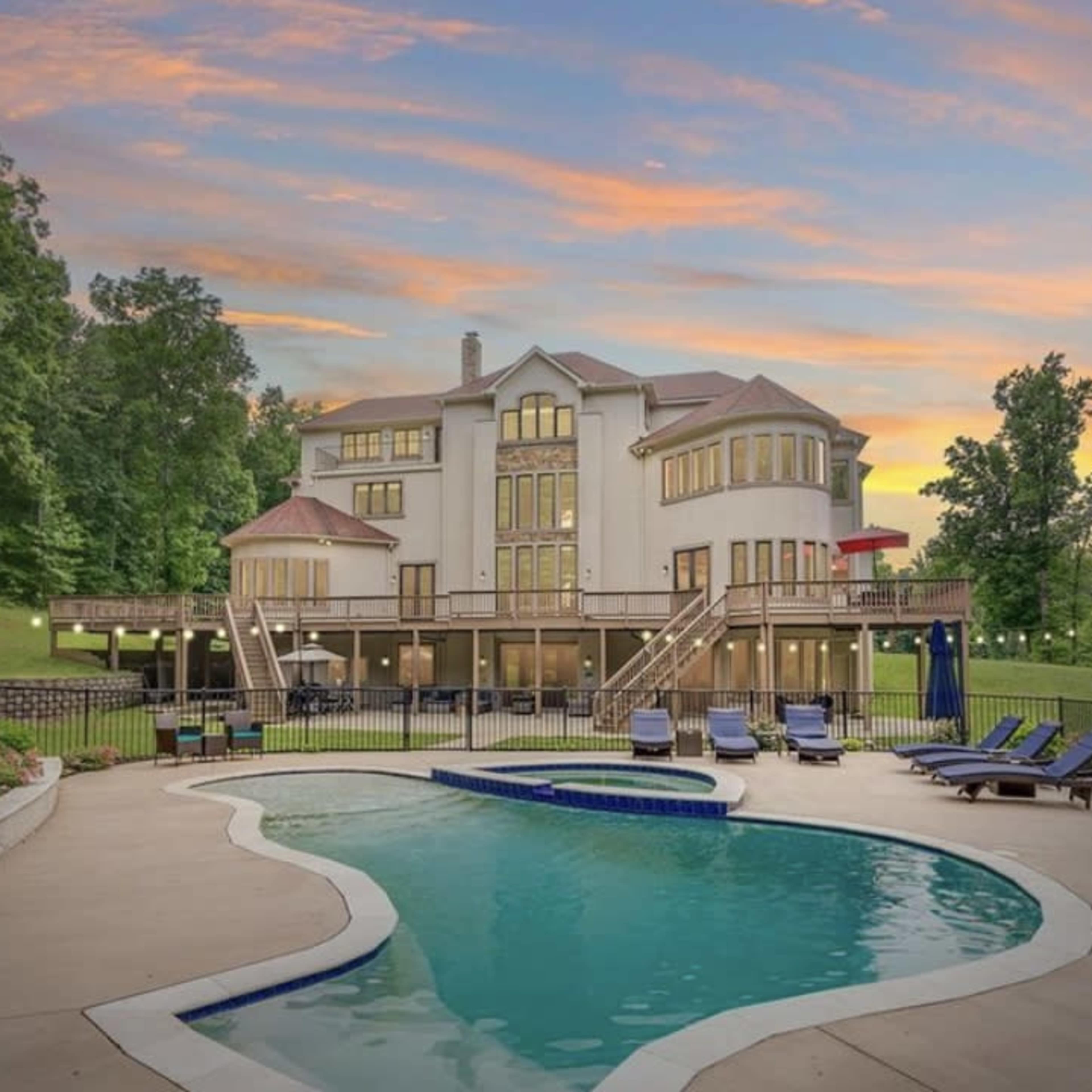 A large house with multiple balconies overlooks a swimming pool surrounded by lounge chairs at sunset.