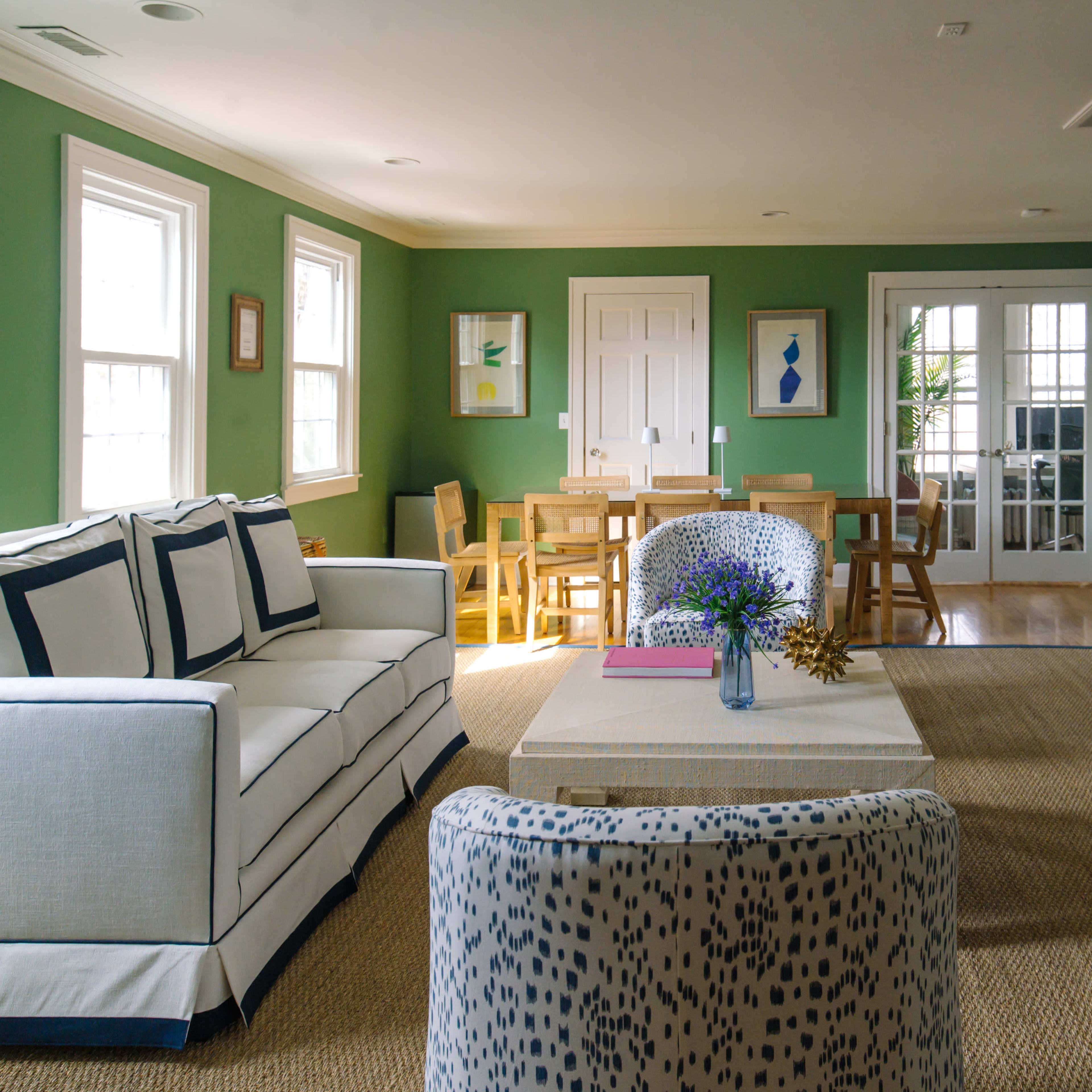 A bright living room features a white couch with navy trim, a coffee table in the center, and a dining area with wooden chairs against green walls.
