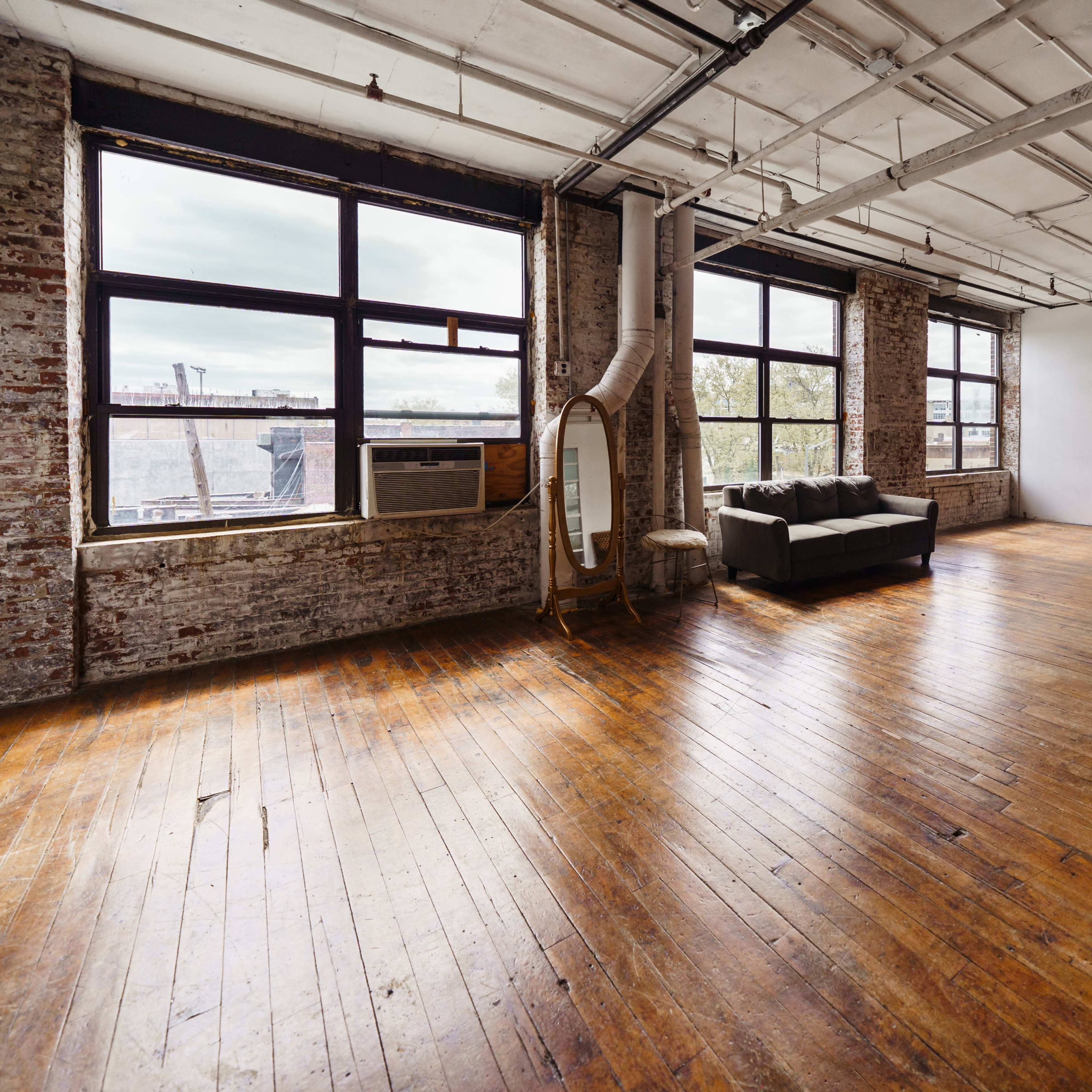 A spacious room with large windows, exposed brick walls, and hardwood flooring, furnished with a sofa and a decorative mirror.