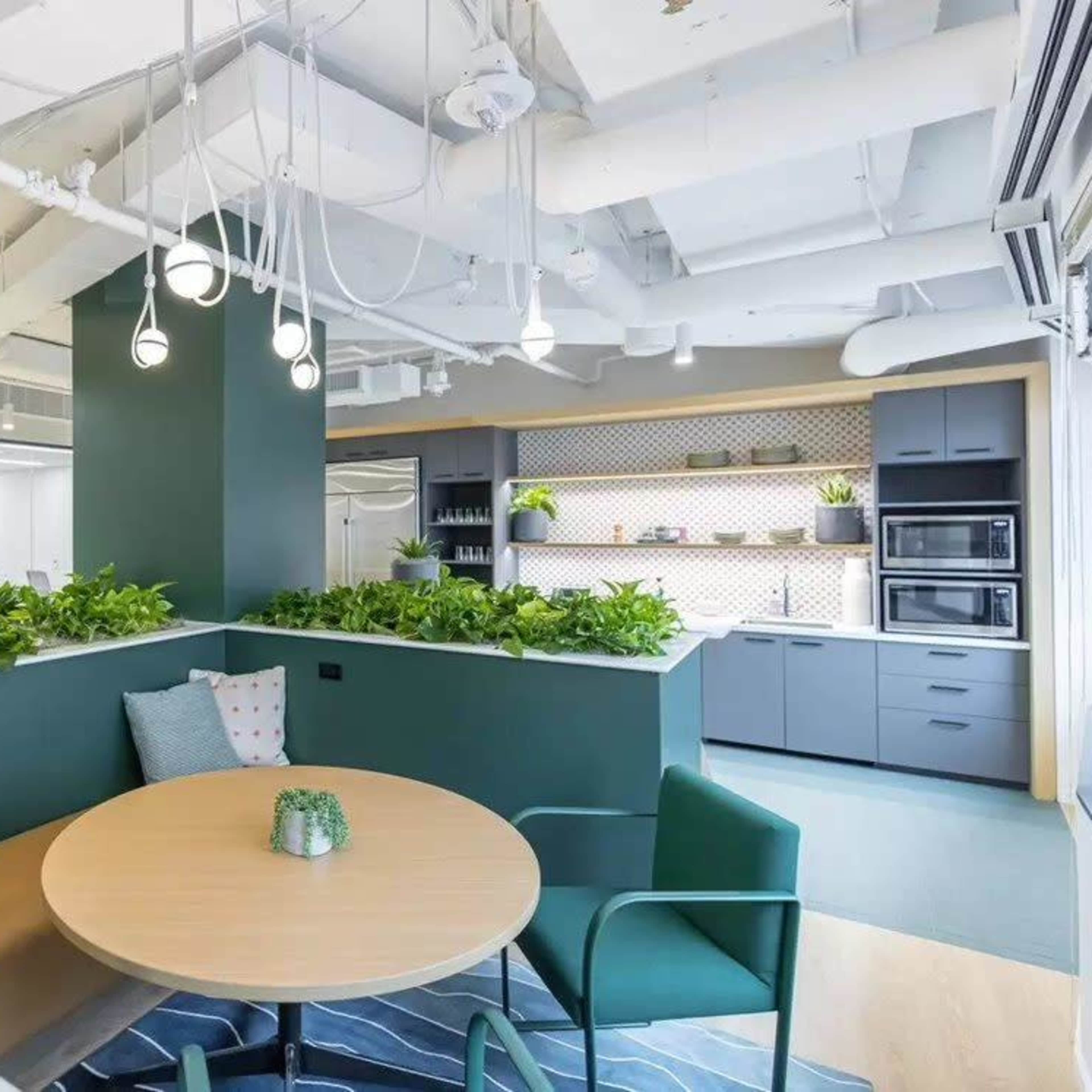 A modern office space with a circular wooden table surrounded by green chairs, flanked by potted plants and a kitchenette in the background.