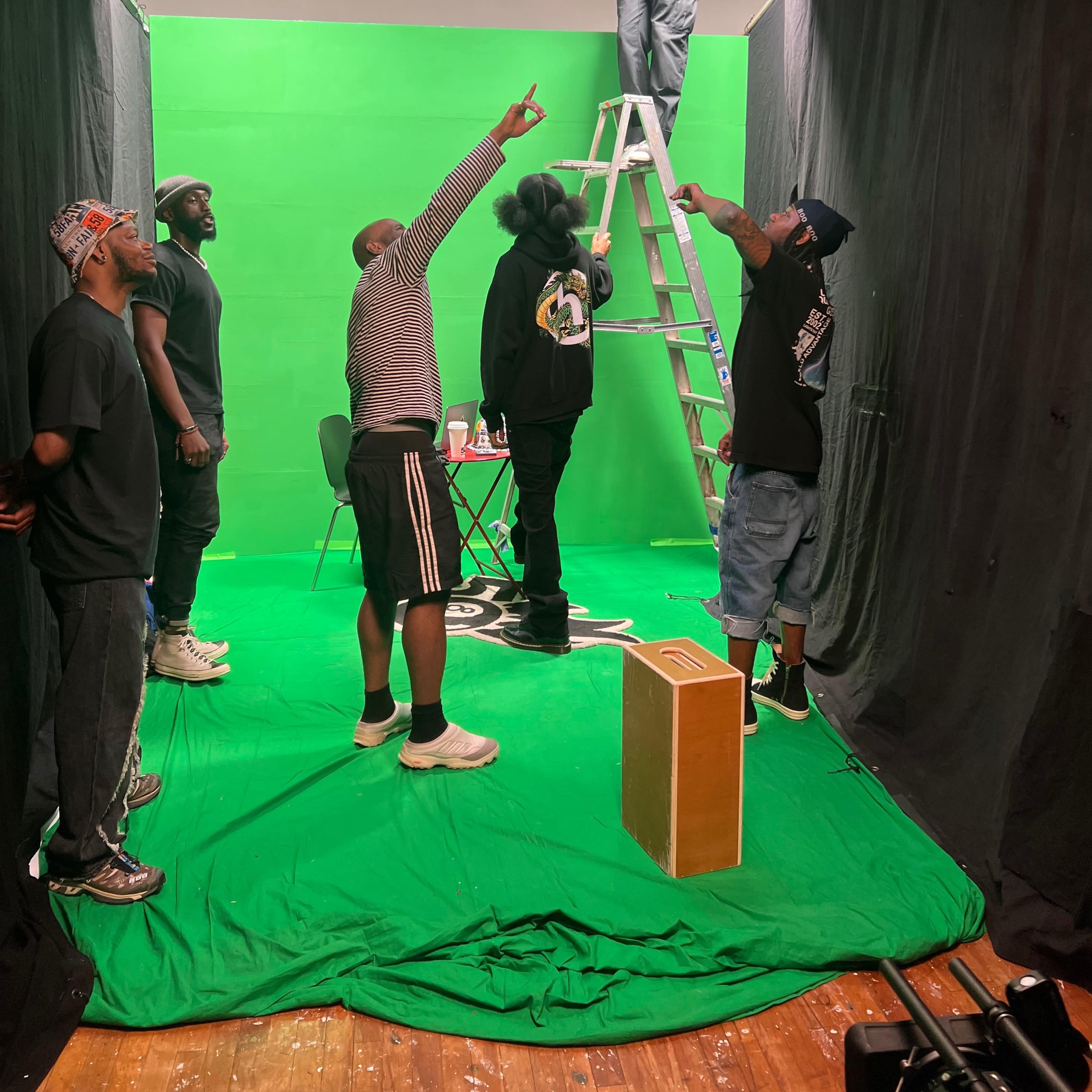 A group of five individuals is directing attention toward a person on a ladder adjusting a light in a green screen studio.