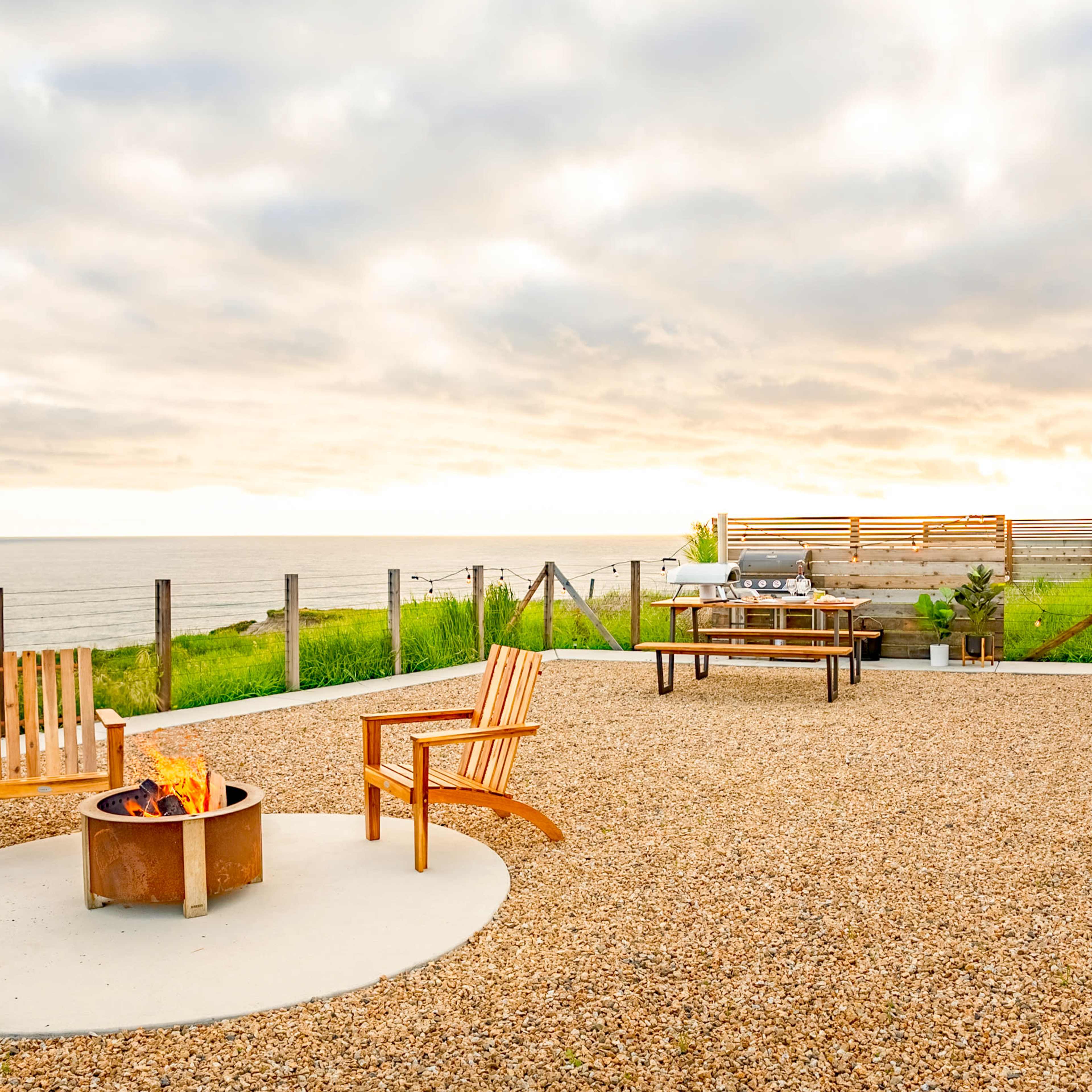 A campground featuring a fire pit surrounded by wooden chairs, gravel ground, and an Airstream trailer overlooking the ocean at sunset.