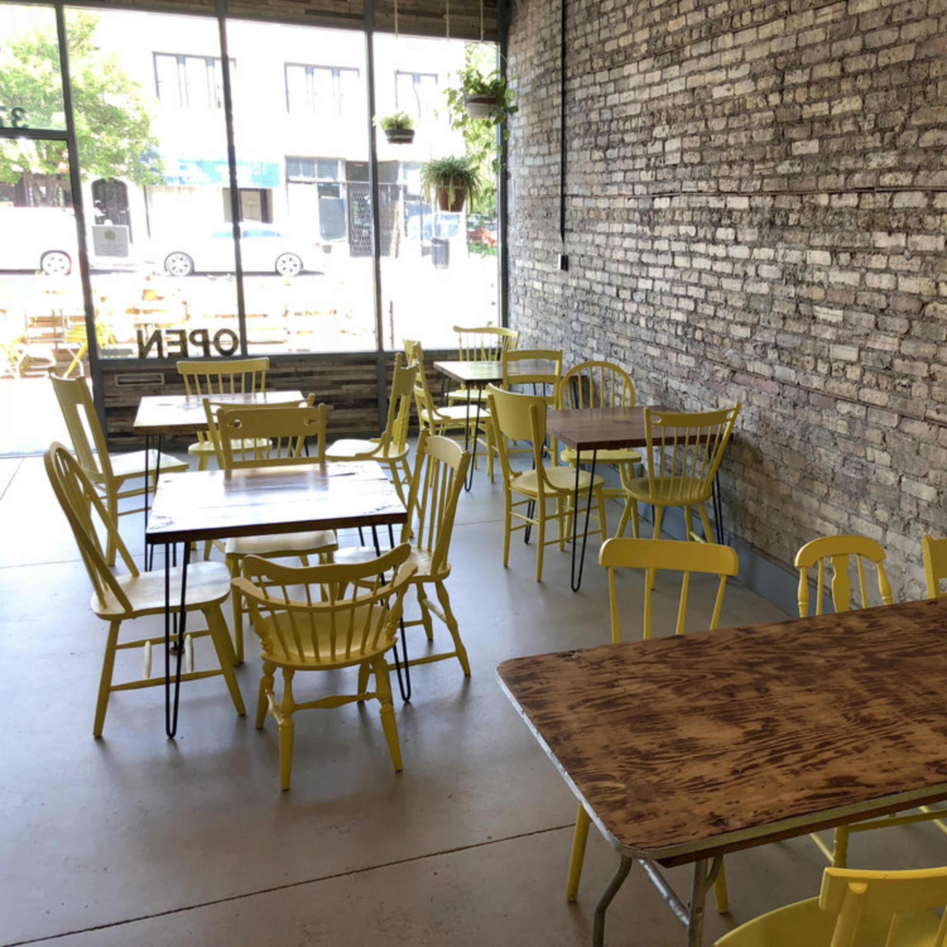 A restaurant interior with yellow chairs and wooden tables arranged on a concrete floor next to large windows.