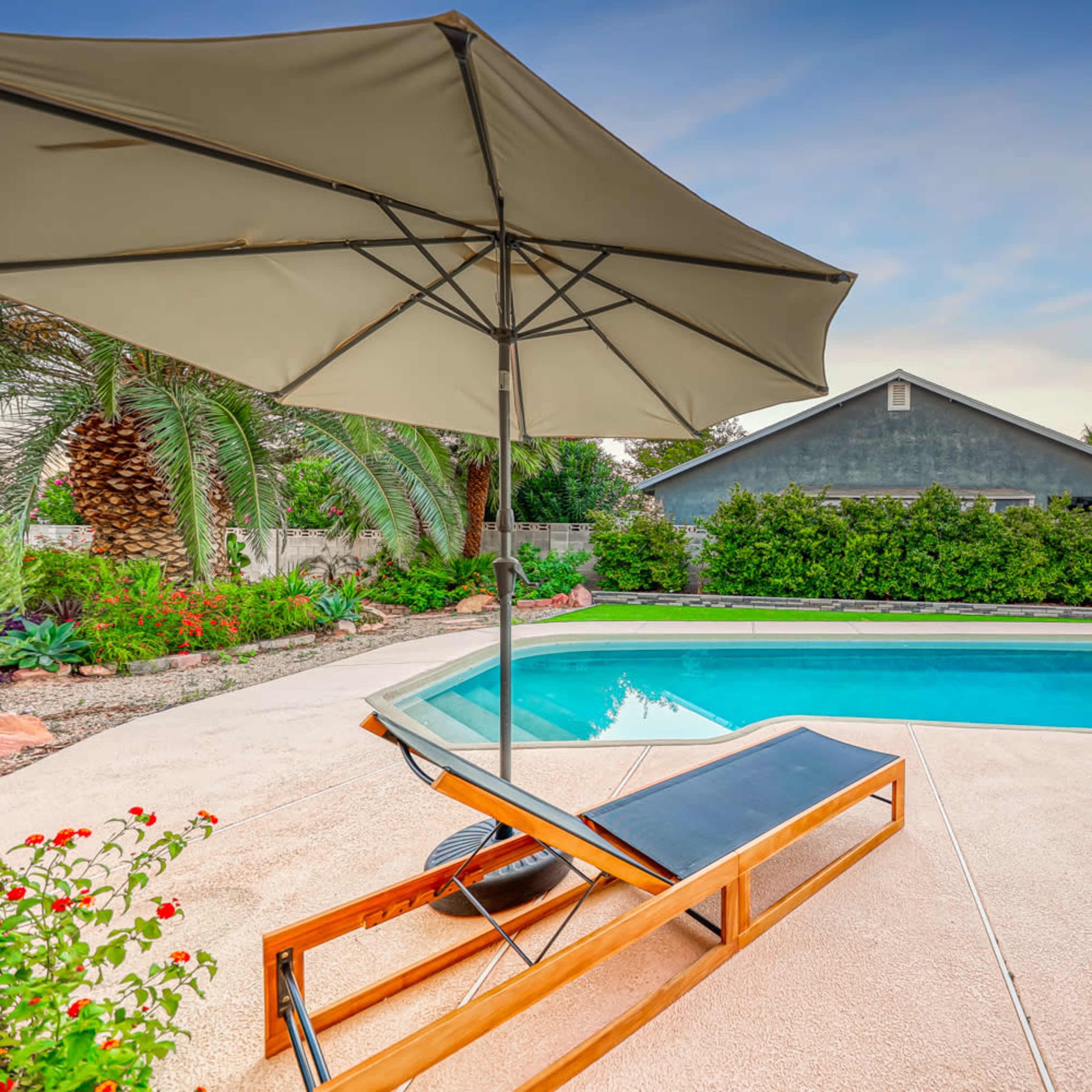A patio with a lounge chair and umbrella overlooks a swimming pool surrounded by greenery and a house in the background.
