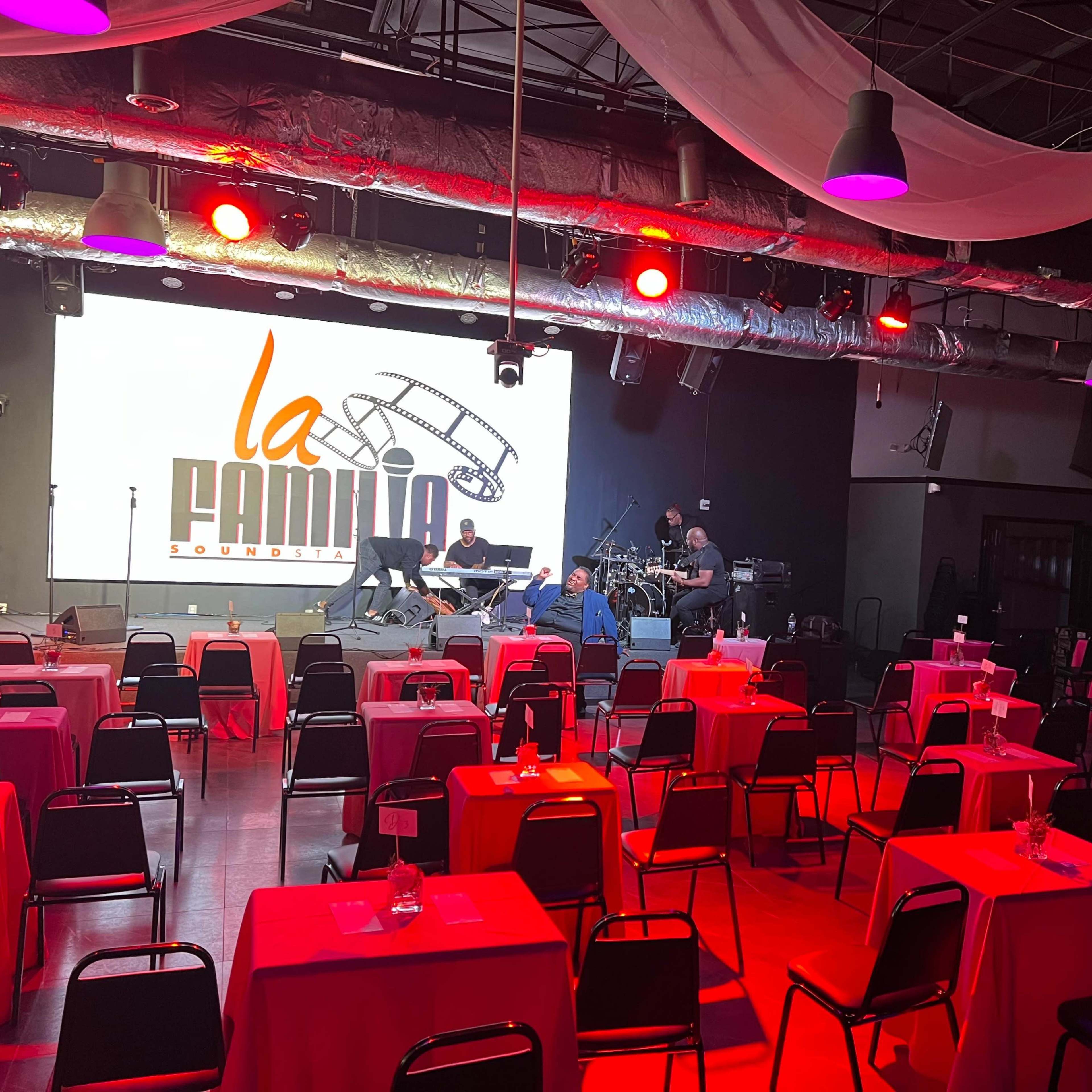 A stage is set up with musical instruments in a dimly lit venue, surrounded by tables covered with red tablecloths.