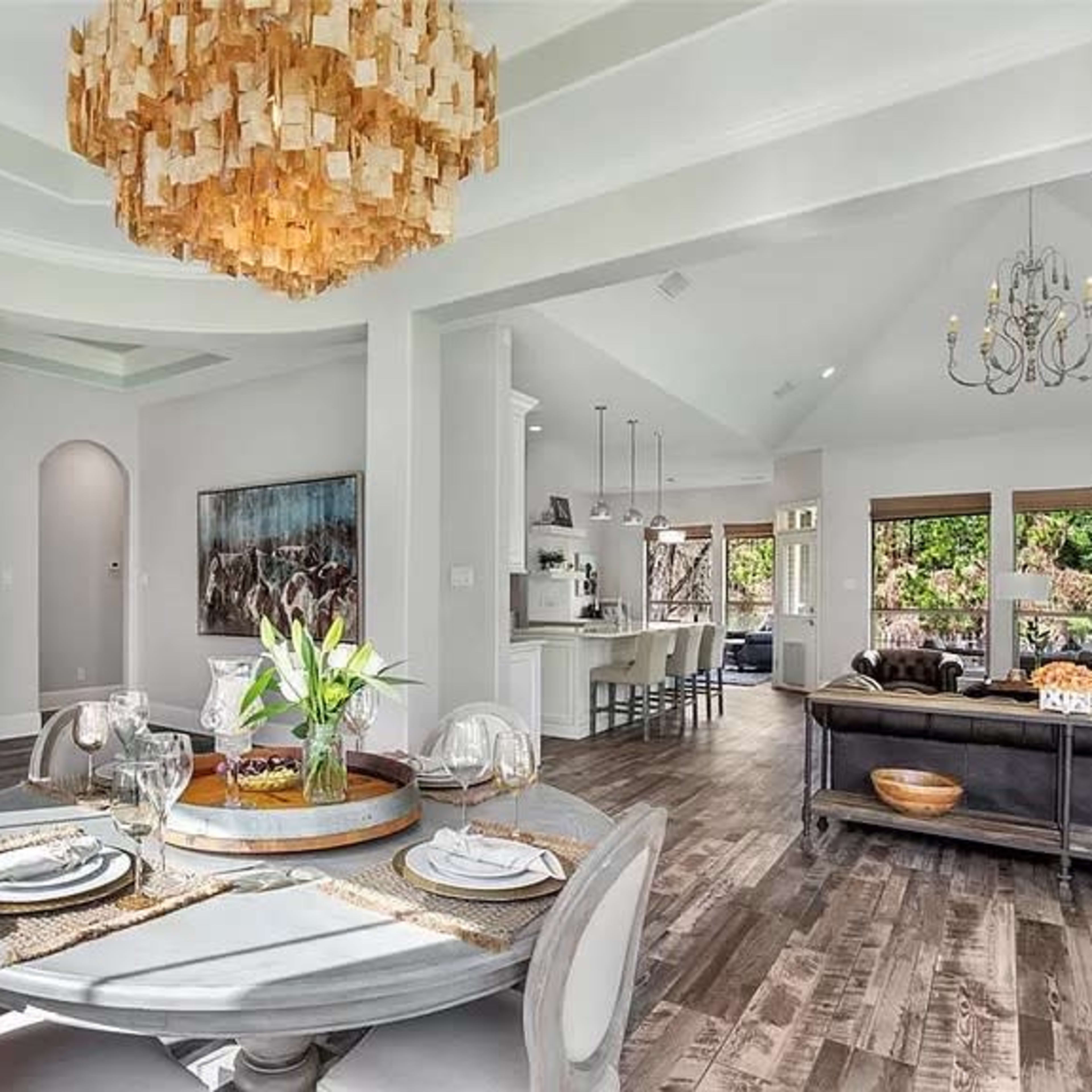 A dining area with a round table set for a meal is visible, adjacent to a spacious living room featuring large windows and a modern chandelier.