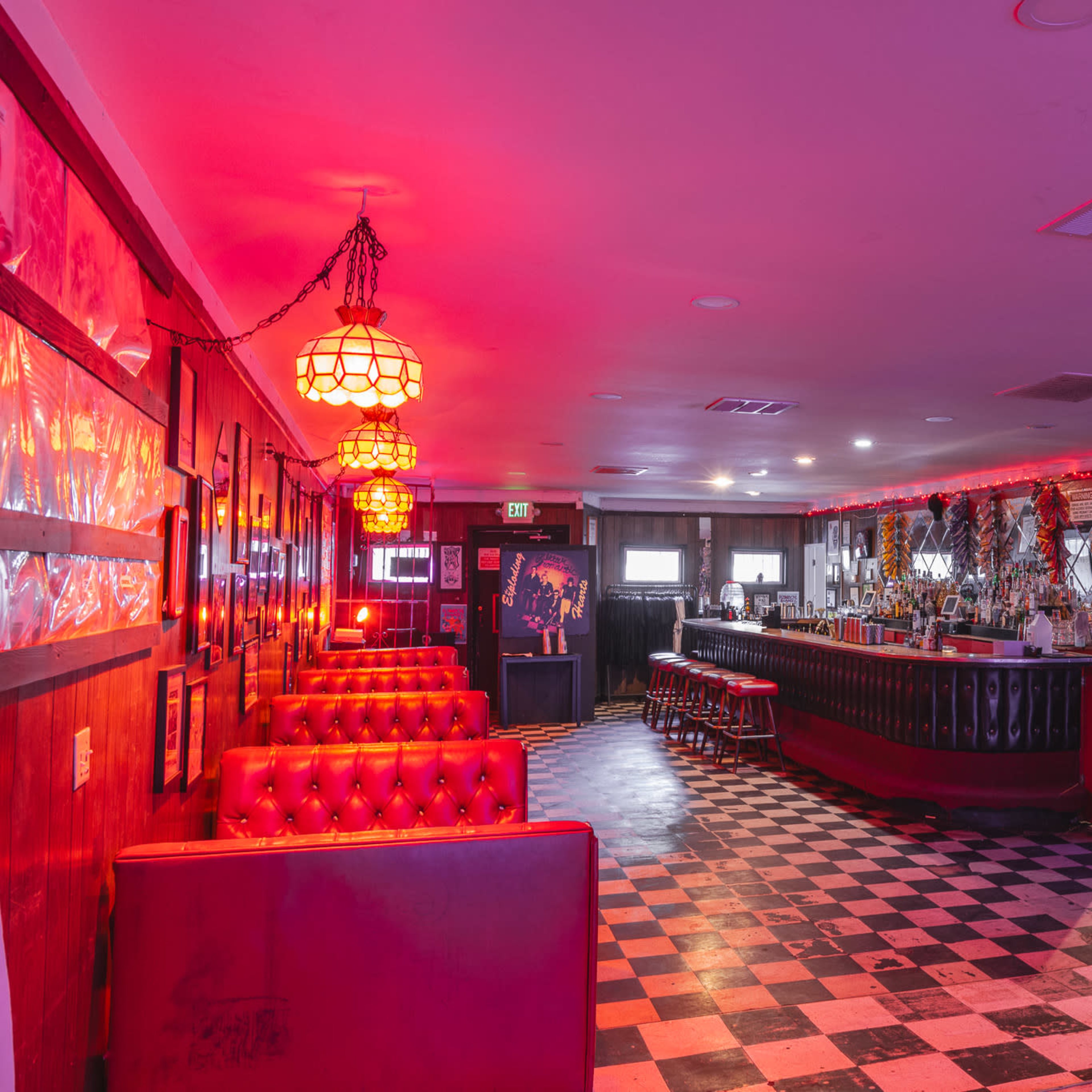 The interior of a bar with red lighting, checkered flooring, and a mix of booth seating and a long bar counter.