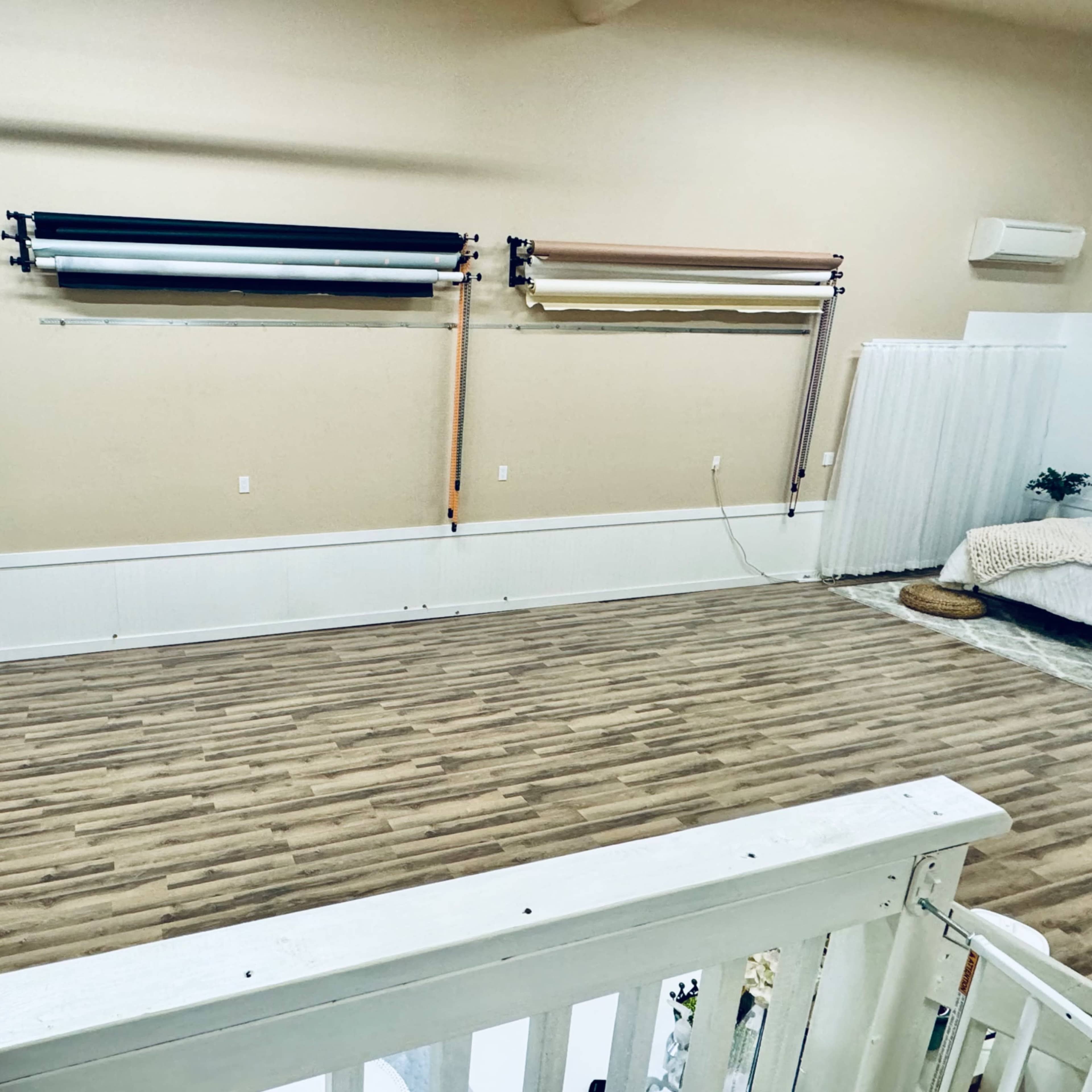 A spacious studio with wooden flooring, equipped with a television, a backdrop system mounted on the wall, and a cozy area with a bed and plants.
