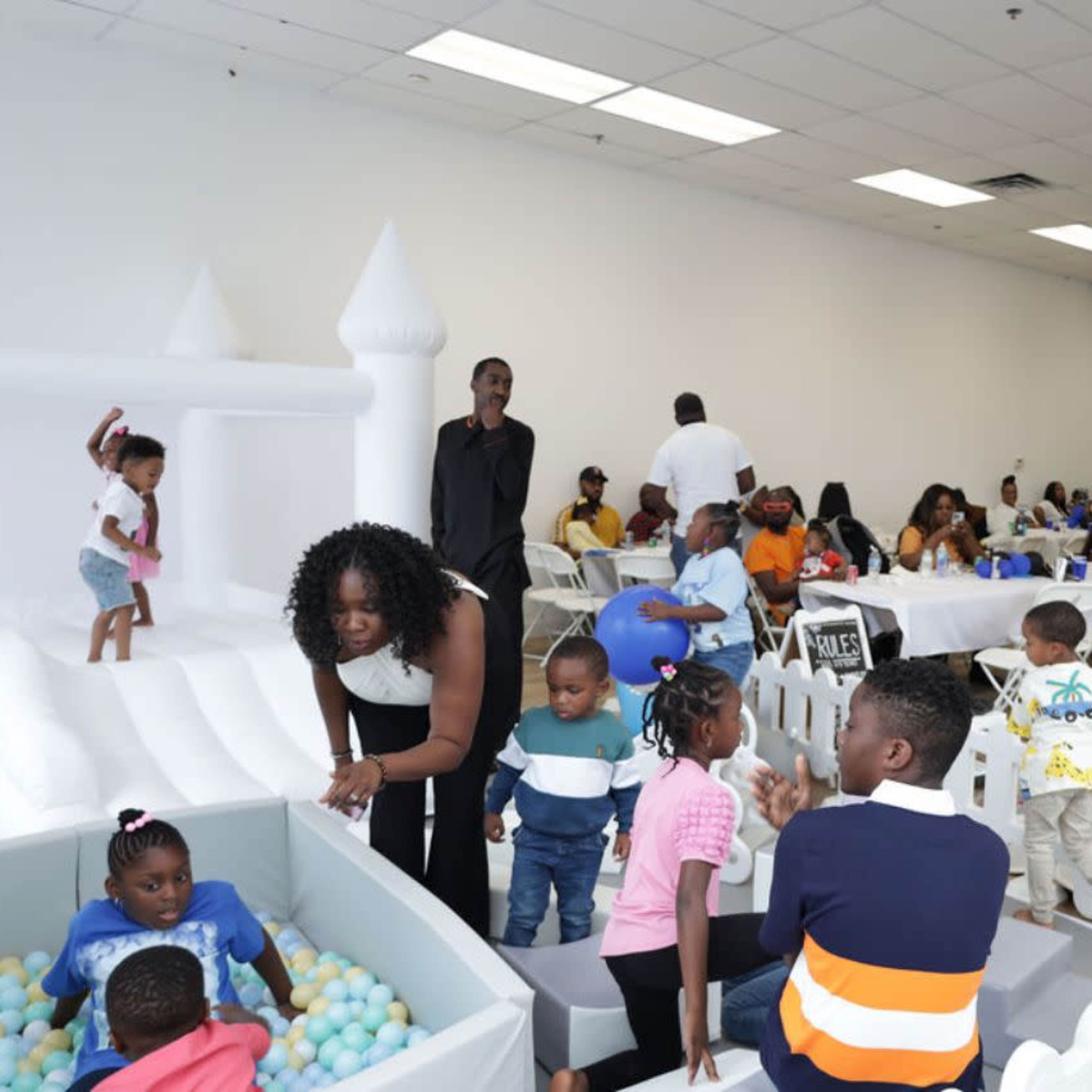 A large indoor event space filled with families, children playing in a ball pit, and tables set for a gathering.