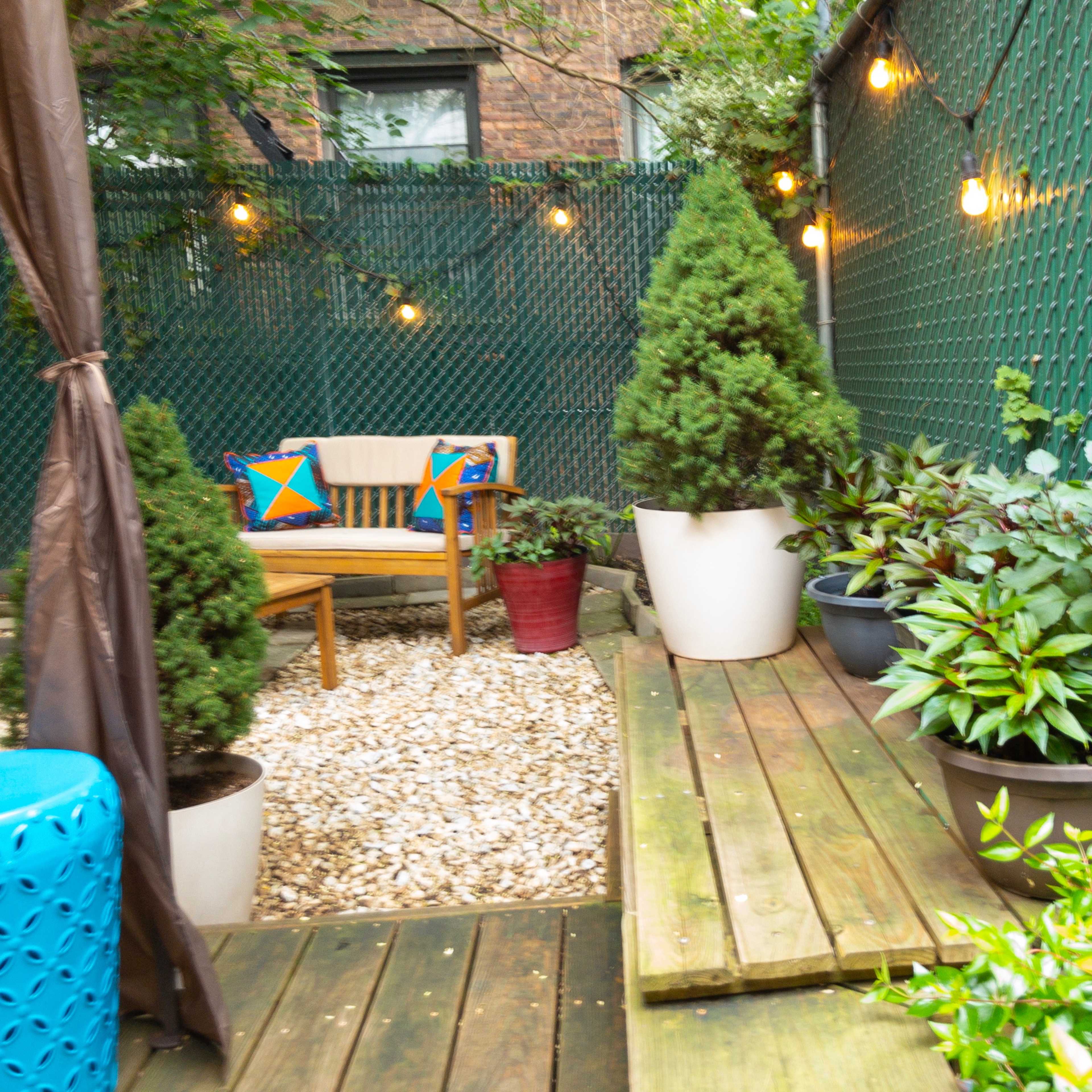 A small outdoor garden features a wooden deck with potted plants, a seating area, and string lights along a green fence.