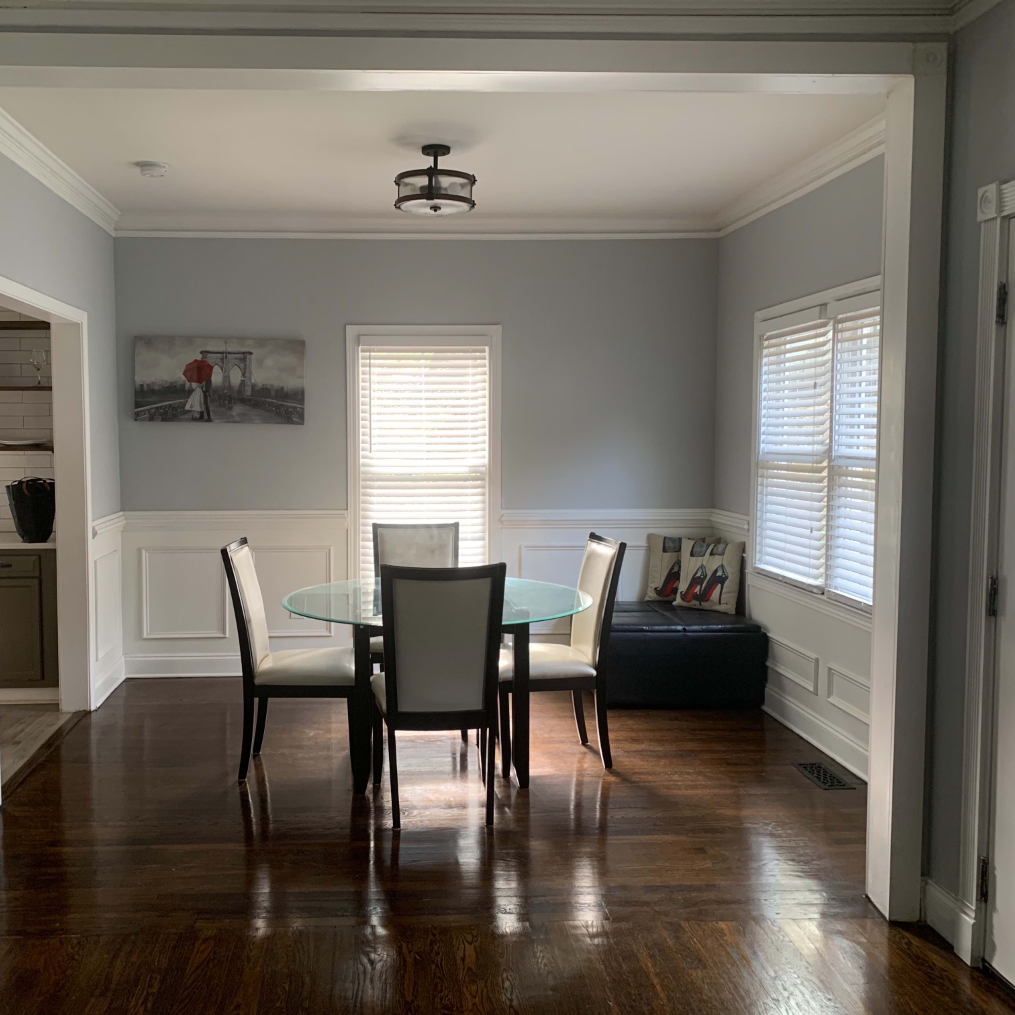 A dining area with a glass table surrounded by four chairs, set against a light blue wall and framed by decorative molding.