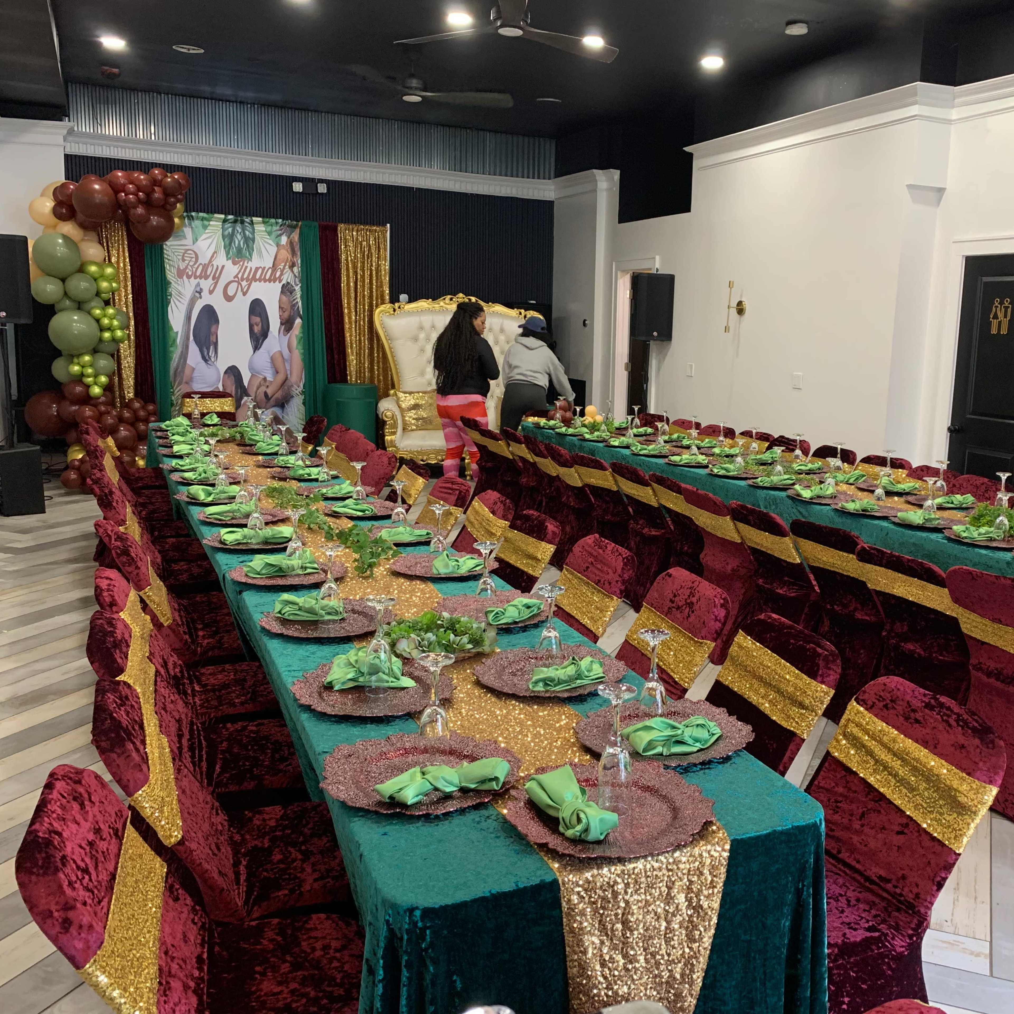 A large event space is set up with two long tables covered in green and maroon tablecloths, adorned with gold accents and neatly arranged place settings.