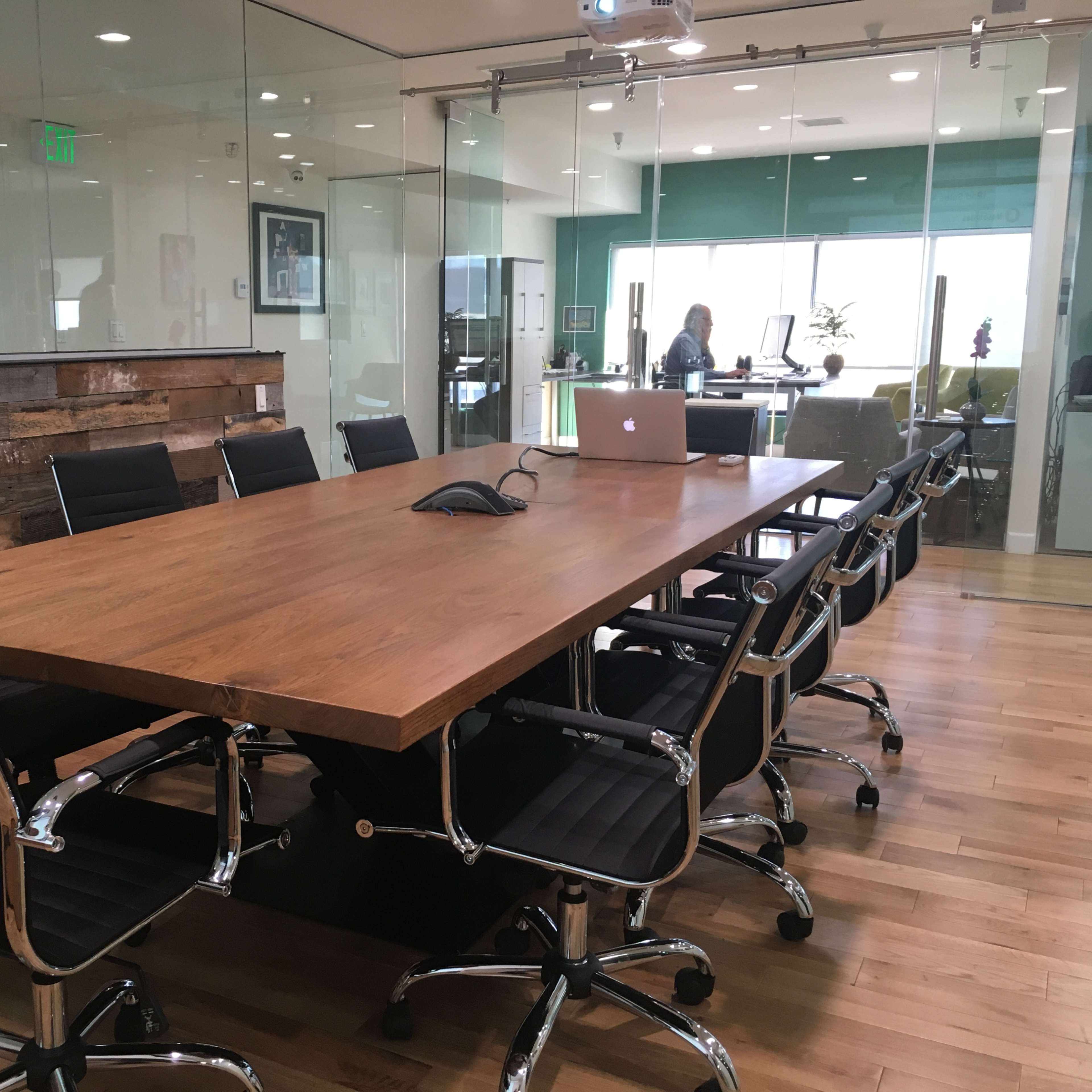 A long wooden conference table surrounded by eight blue upholstered chairs is set in a well-decorated meeting room with potted plants and a wooden accent wall.