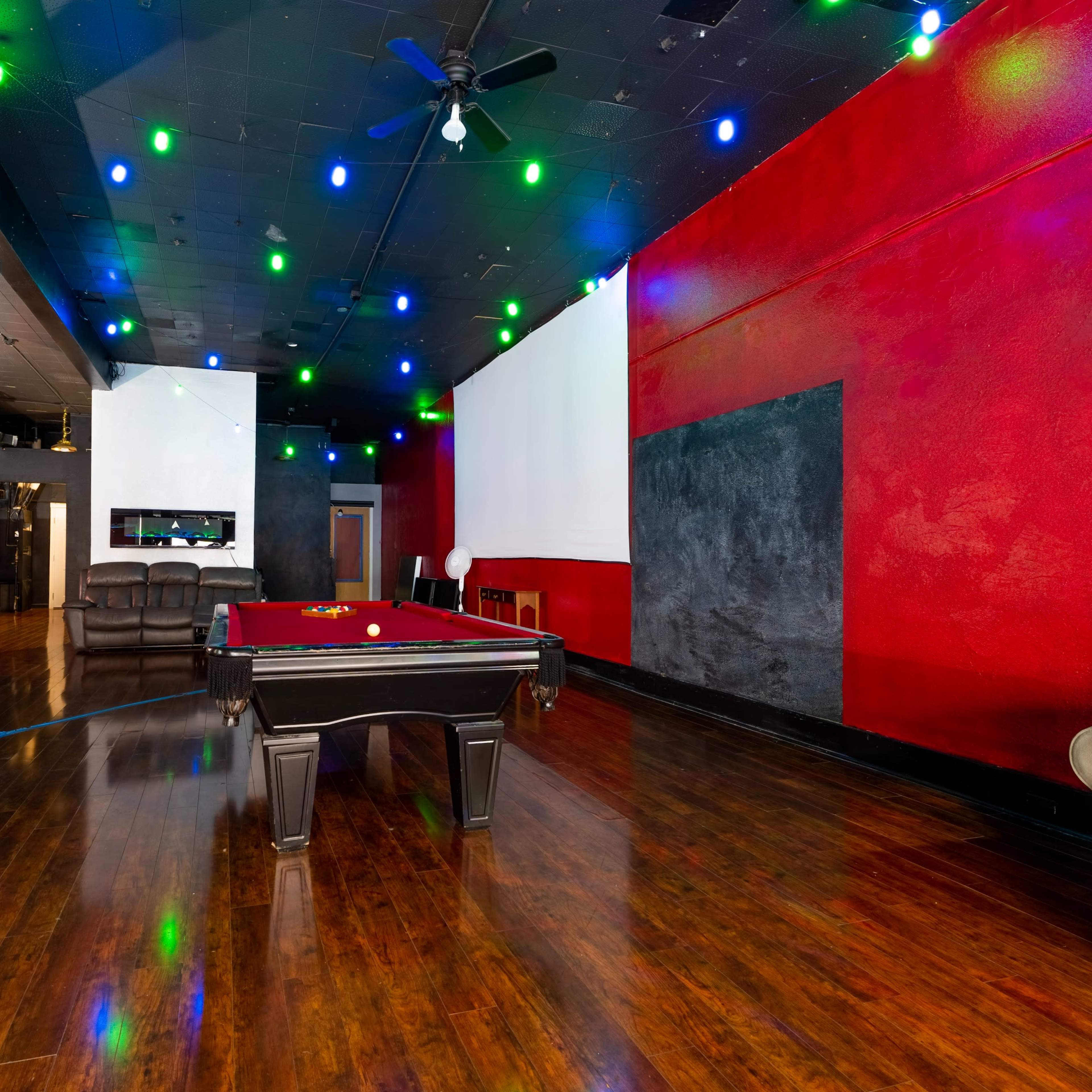 A game room with a billiard table in the center, surrounded by red walls, hardwood floors, and colorful ceiling lights.