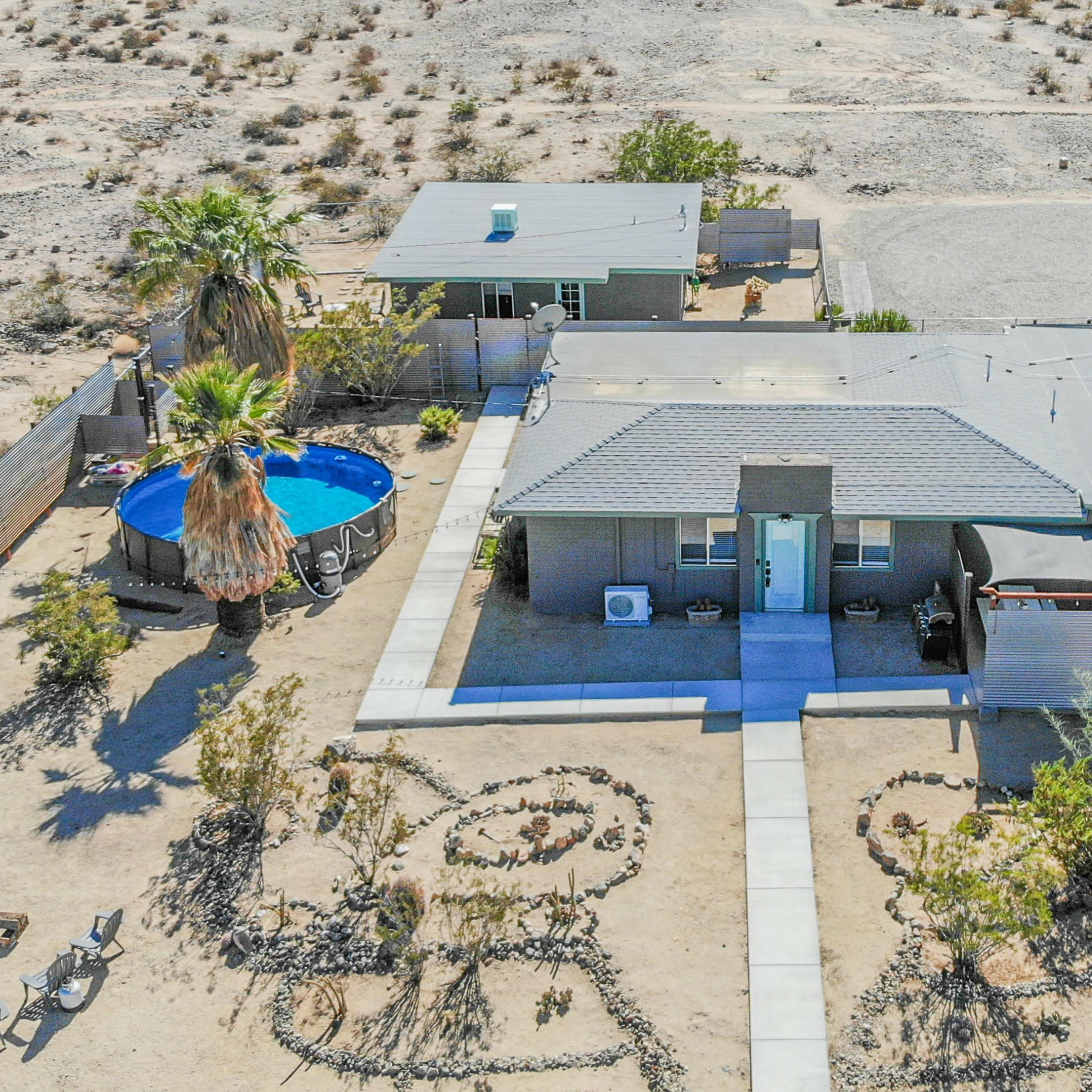 An aerial view of a modest home surrounded by desert landscaping, with a swimming pool, two additional structures, and a neatly laid pathway.