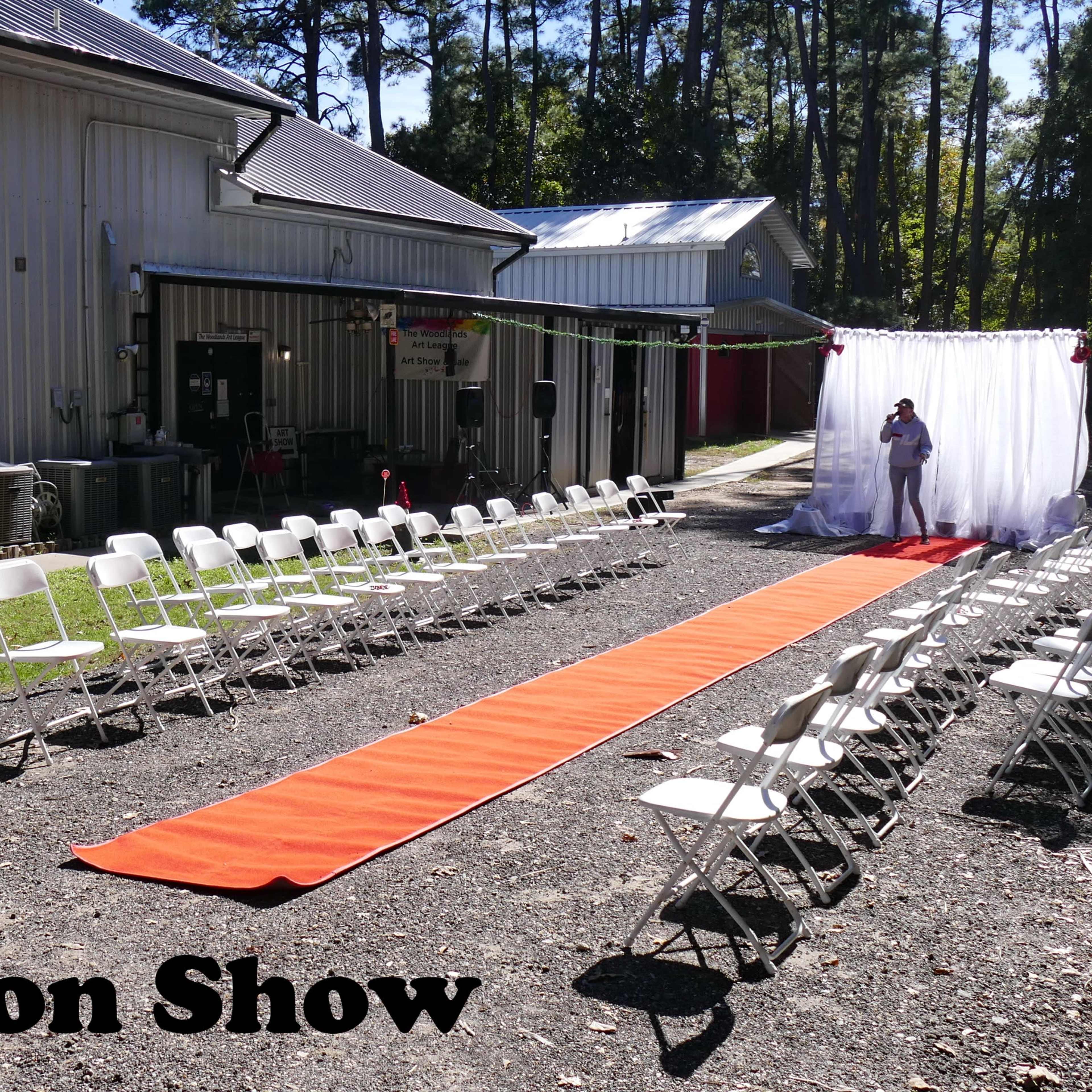 A fashion show setup features a runway with an orange carpet, flanked by rows of white chairs, in an outdoor area behind a building.