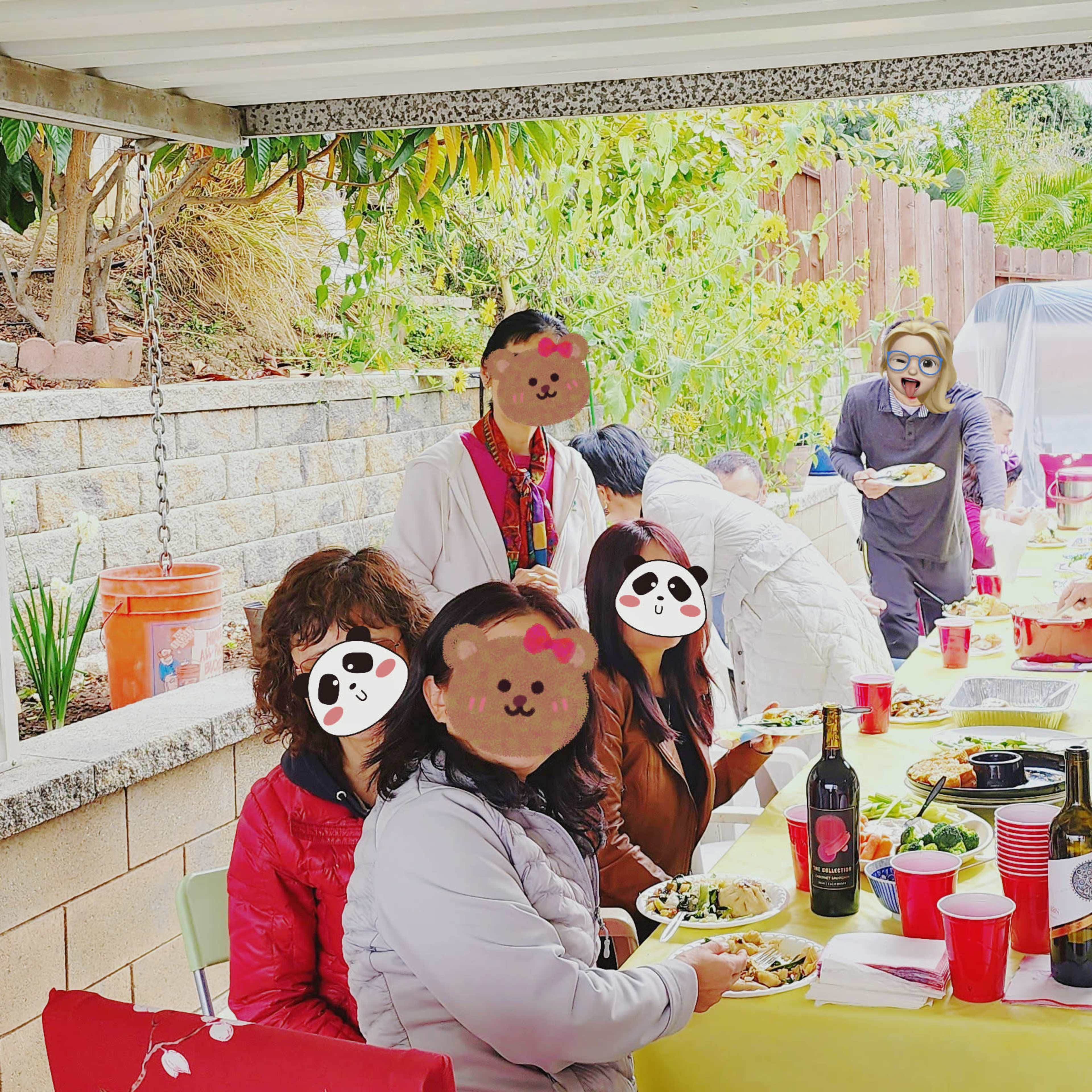 A group of people gathers around a long table under a patio, sharing food and drinks at a casual outdoor gathering.