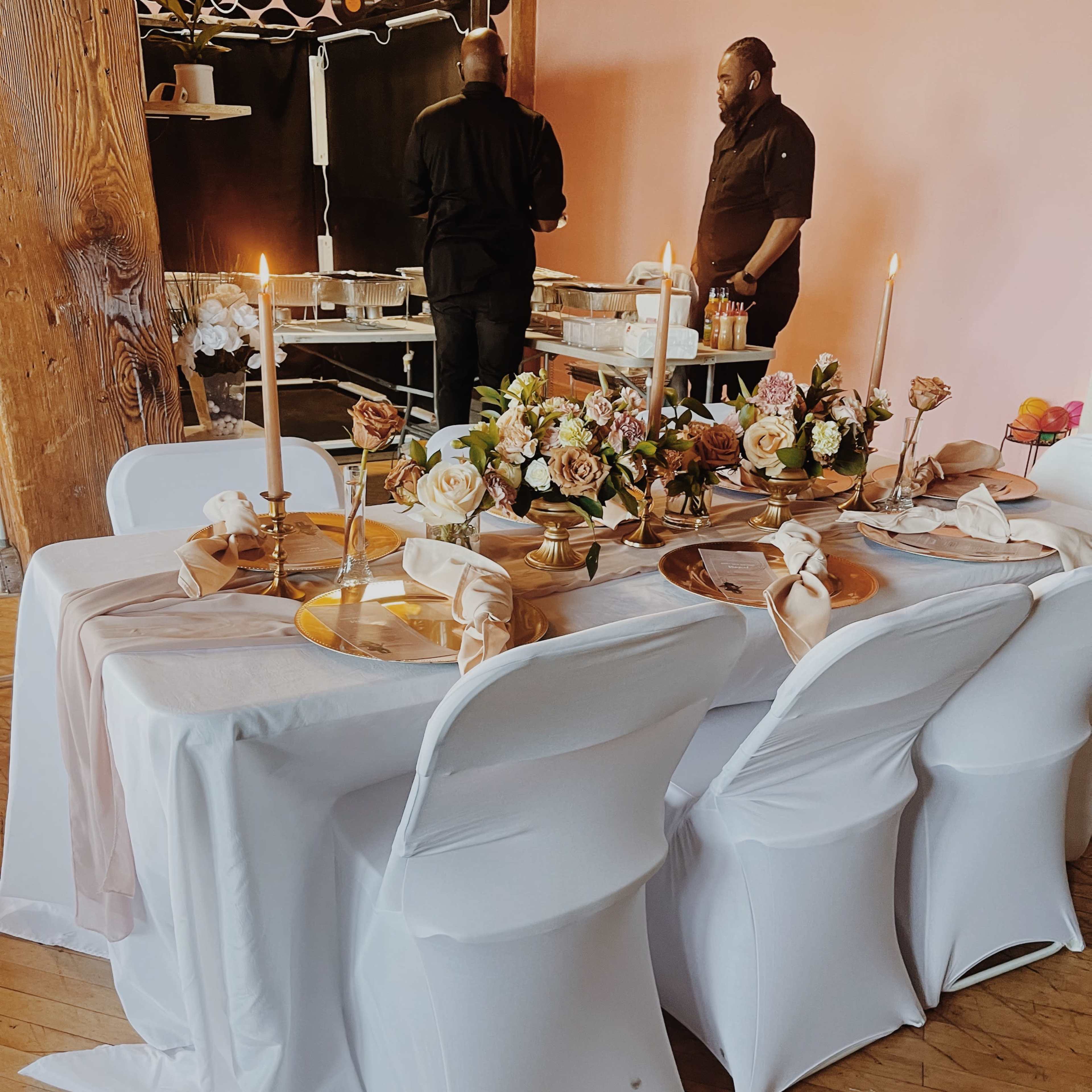 A table adorned with a floral centerpiece, candles, and elegant place settings is set up in a dining area with two people preparing food in the background.
