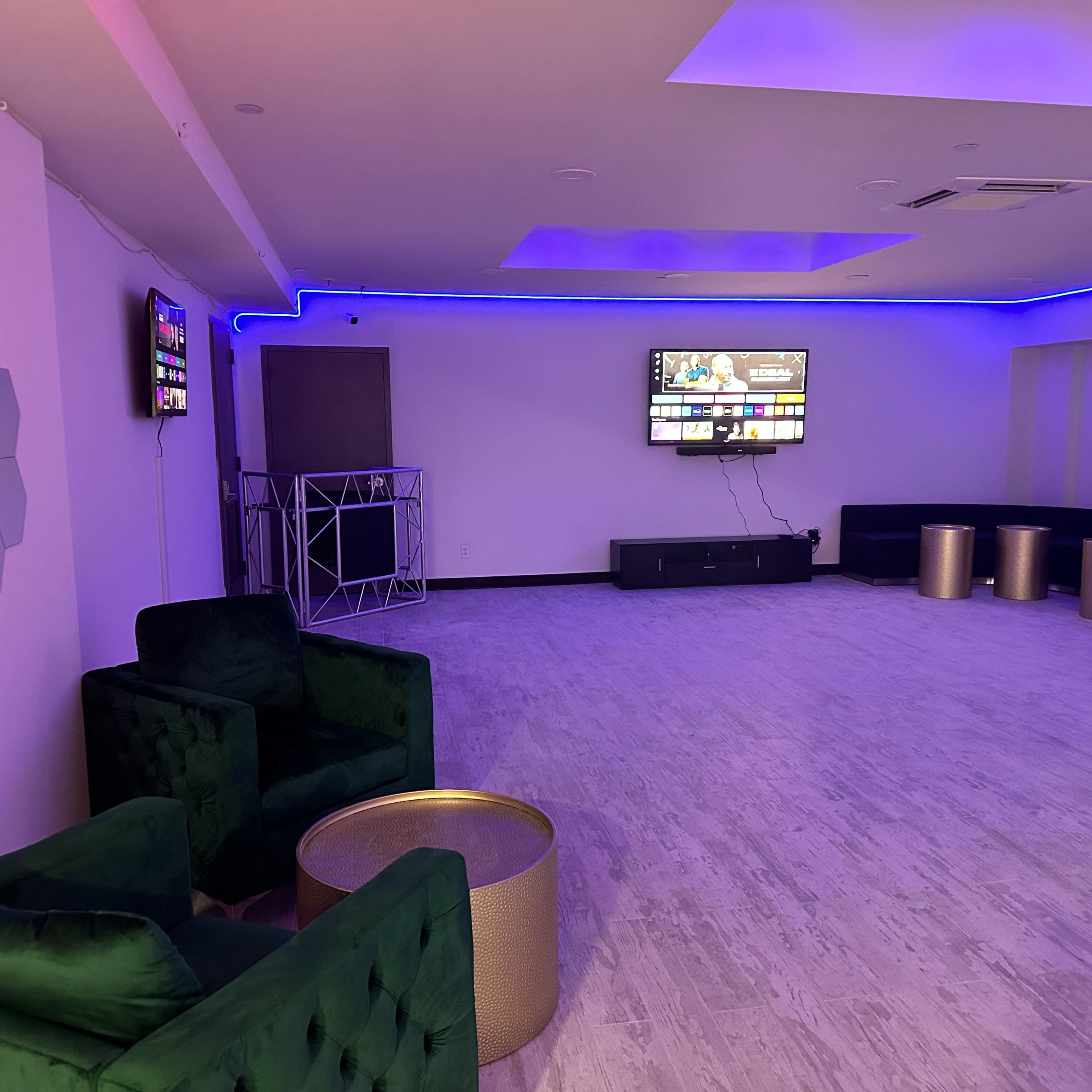 A modern, minimally furnished room features green velvet chairs, a gold table, and a wall-mounted television, with blue LED lighting along the ceiling.