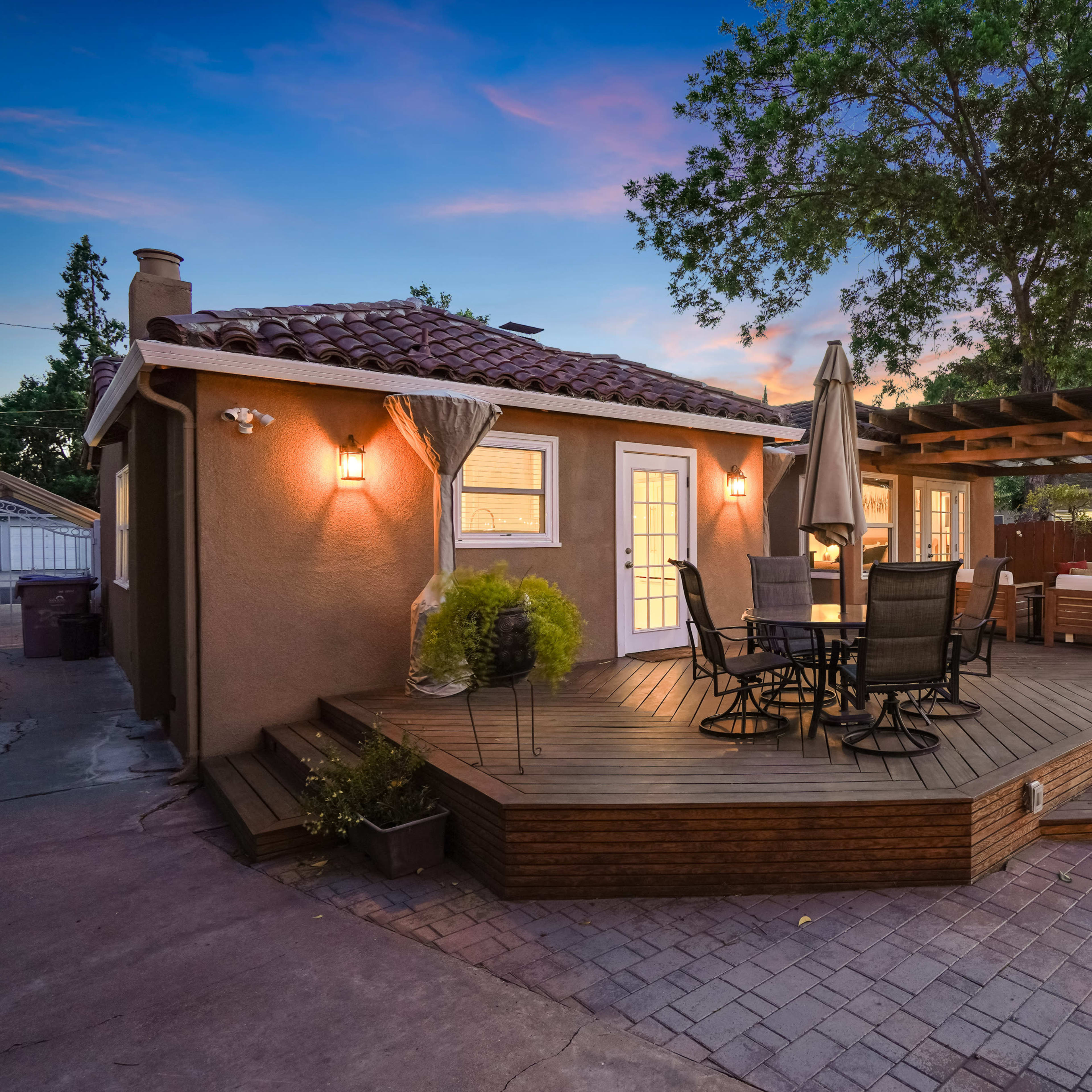 A wooden deck with a dining table and chairs is connected to a house, surrounded by greenery and outdoor furniture, set against a twilight sky.