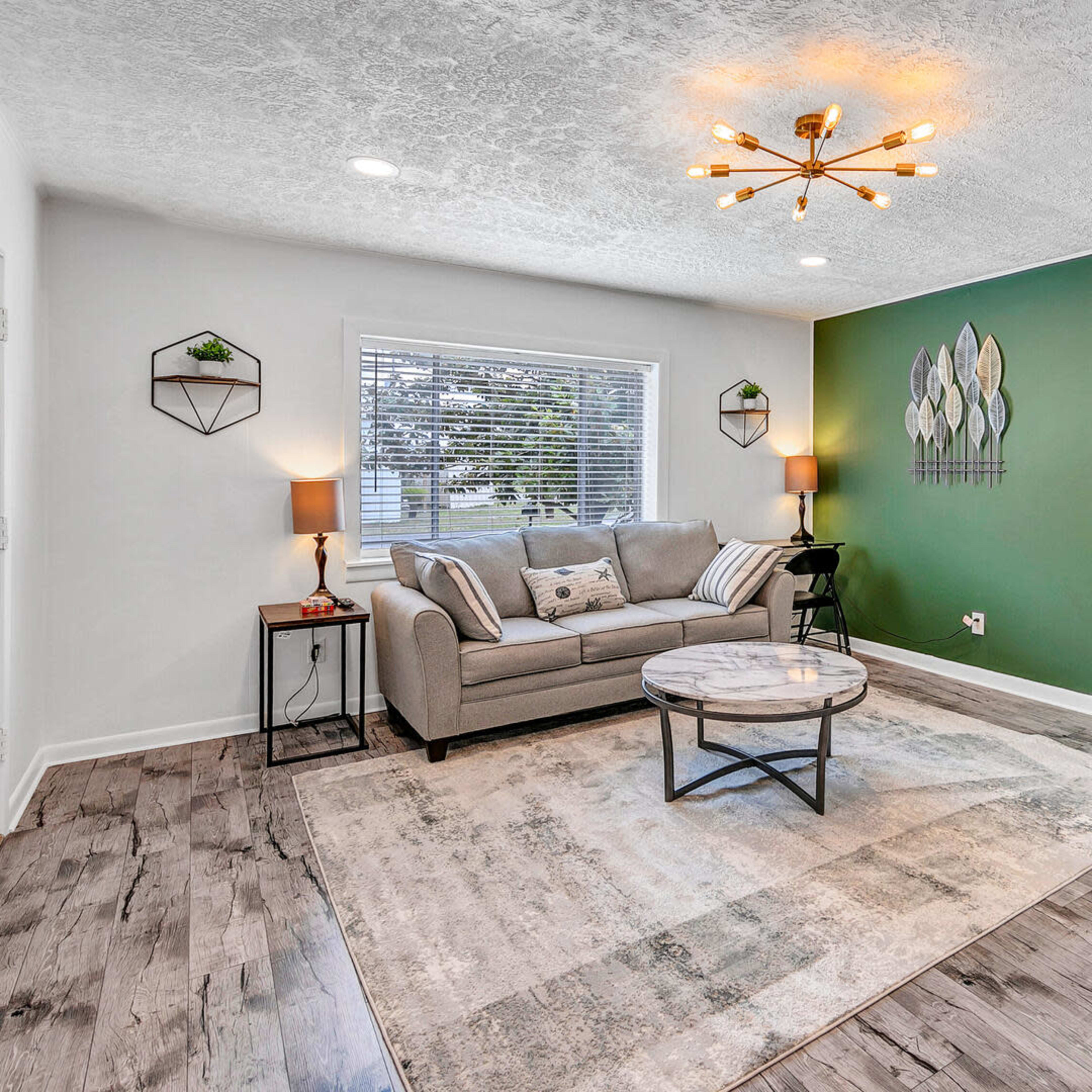 A modern living room with a gray sofa, a round coffee table, and green accent walls, leading to a kitchen area in the background.