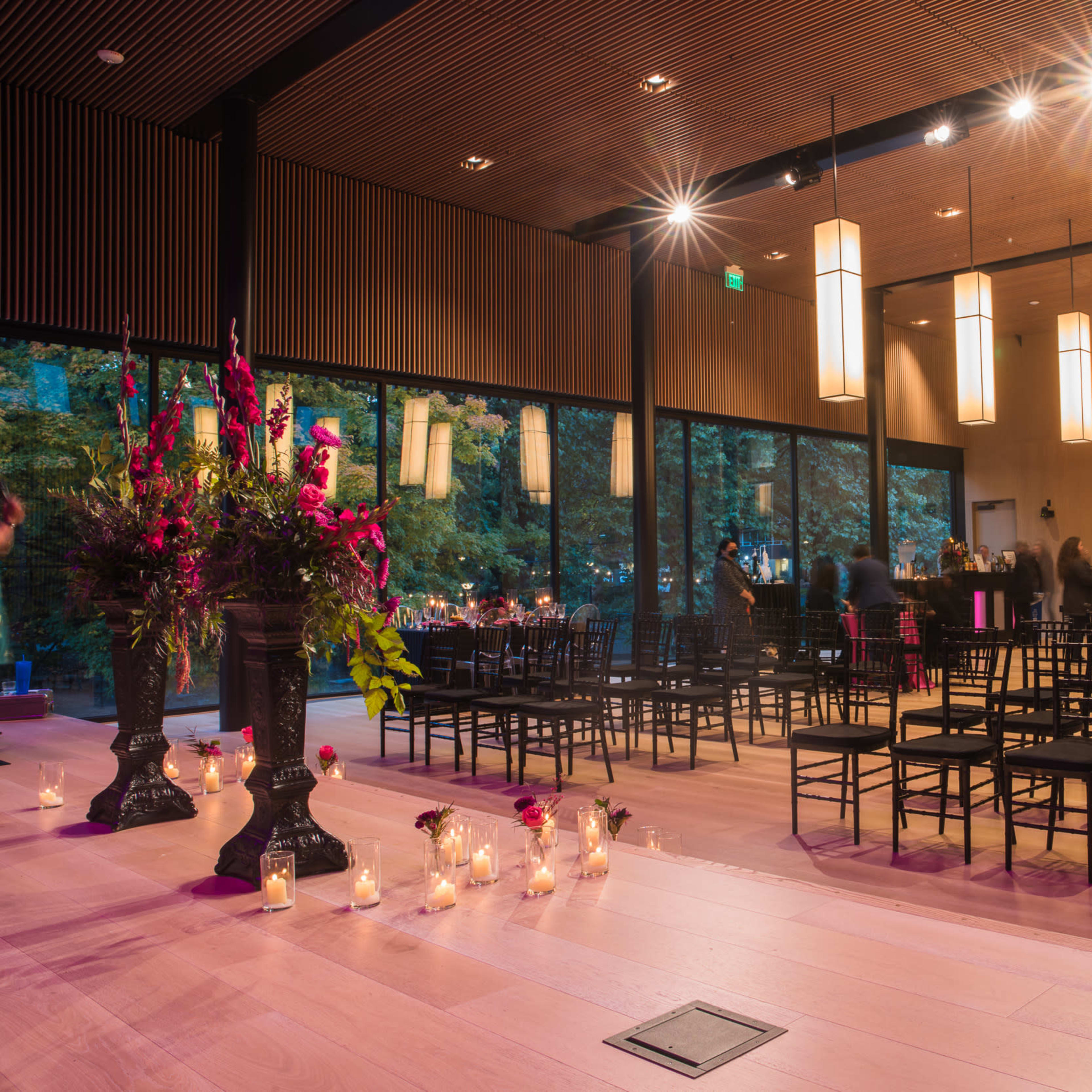 An indoor venue with rows of black chairs, a floral arrangement, and soft lighting from hanging lamps, creating a space set up for an event.