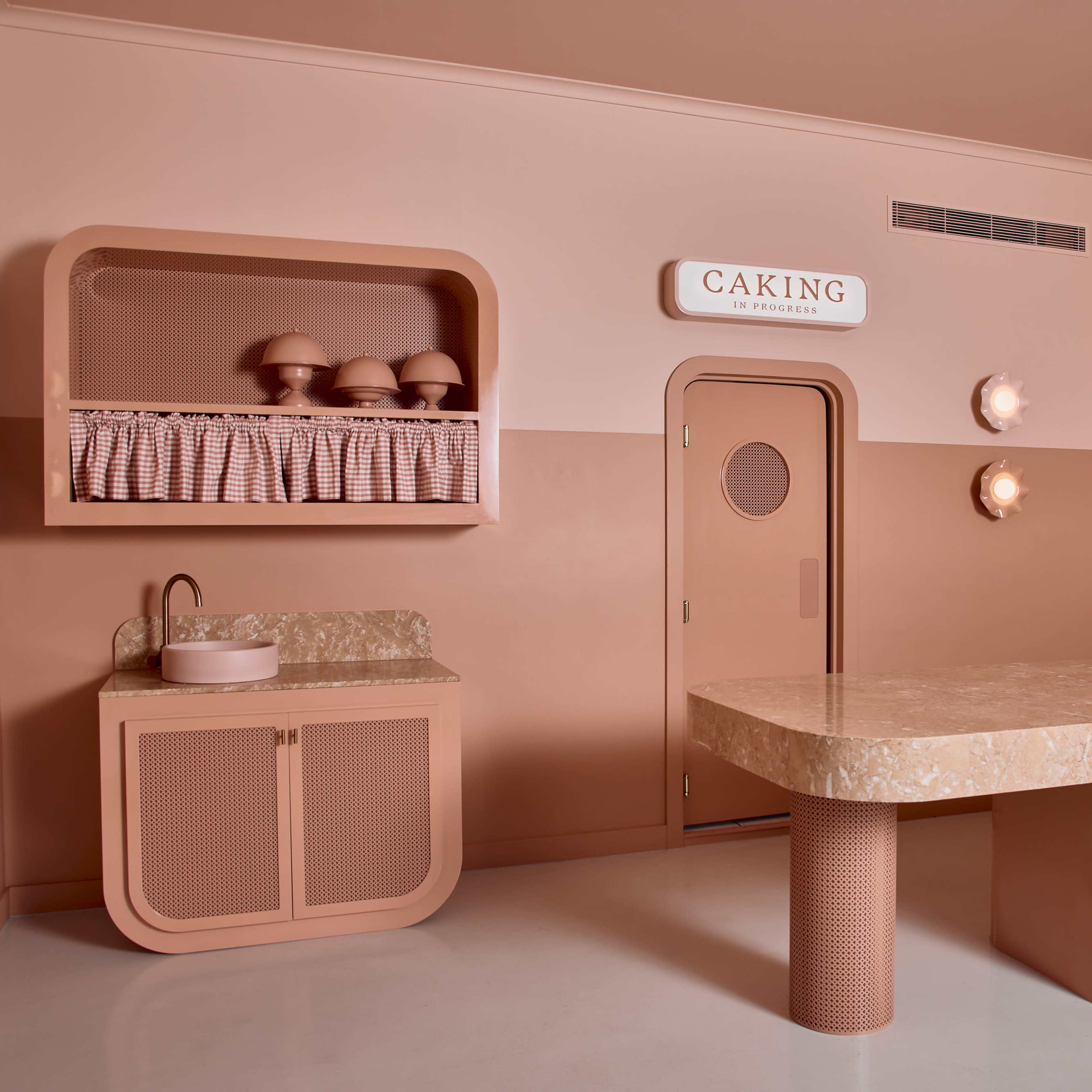 A minimalist interior space with beige and peach walls, featuring a large marble-topped table, a washing area, and a door labeled "CAKING."