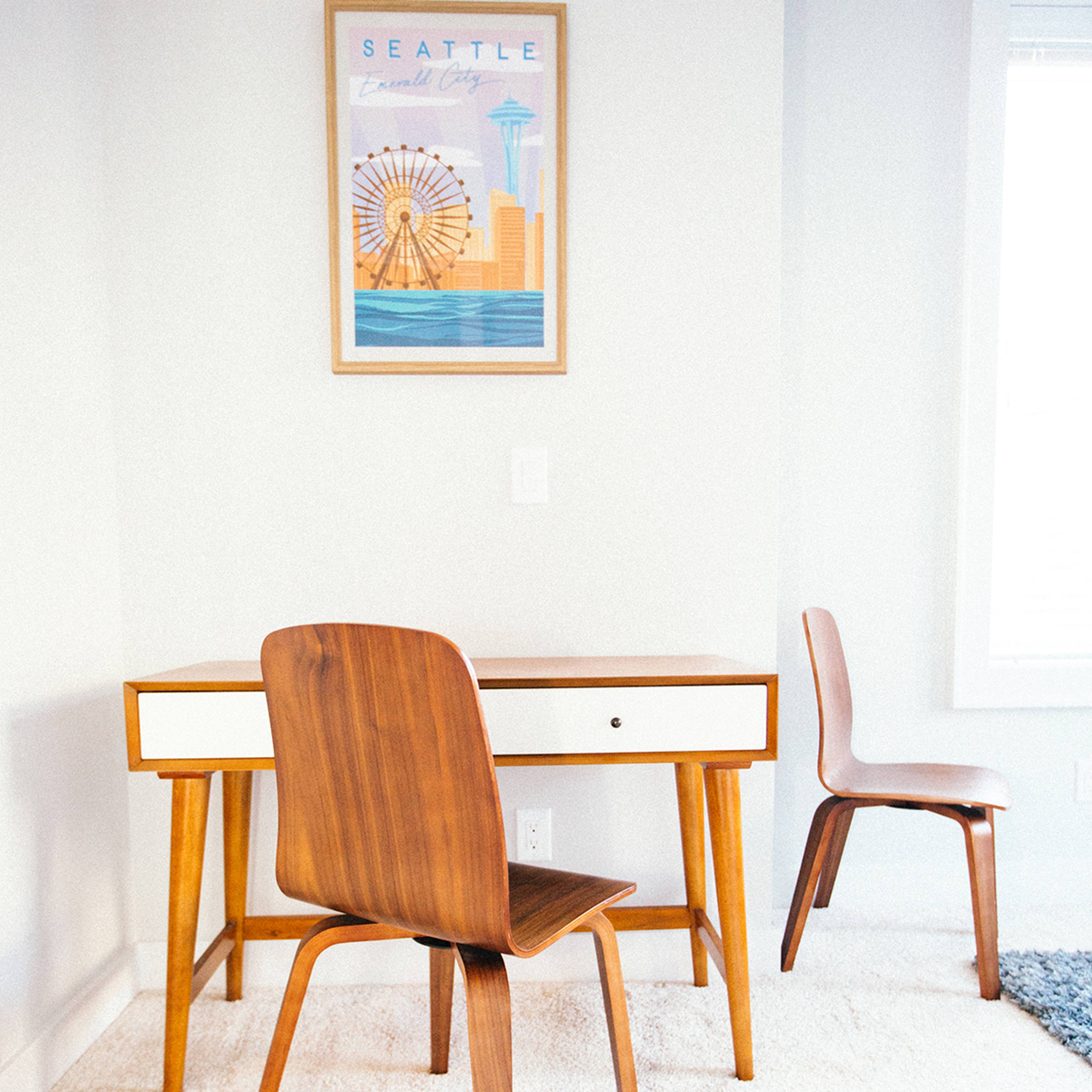 A wooden desk and two chairs are situated in a bright room with a Seattle-themed poster hanging on the wall.