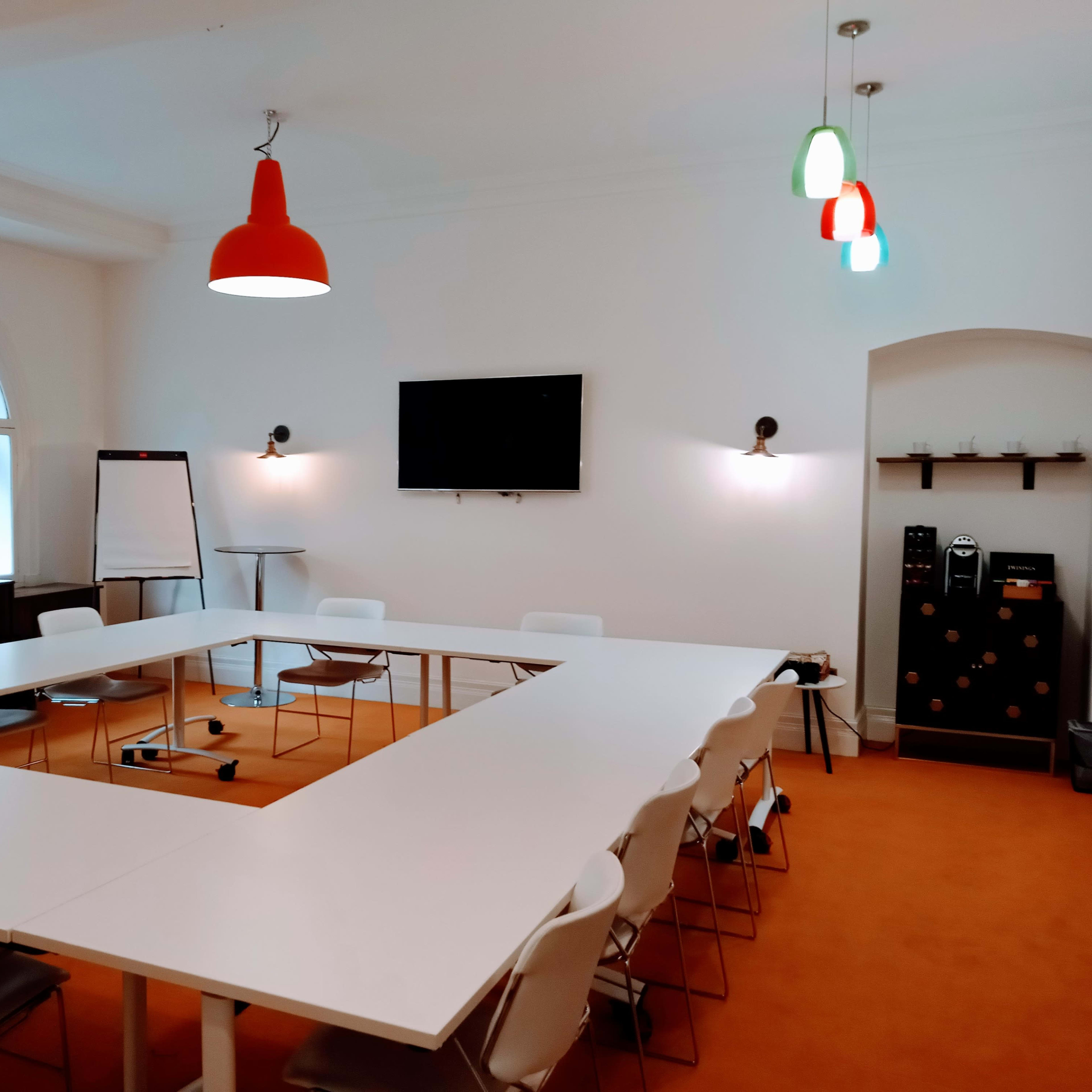 A conference room features a white U-shaped table surrounded by chairs, a television mounted on the wall, and a window letting in natural light.