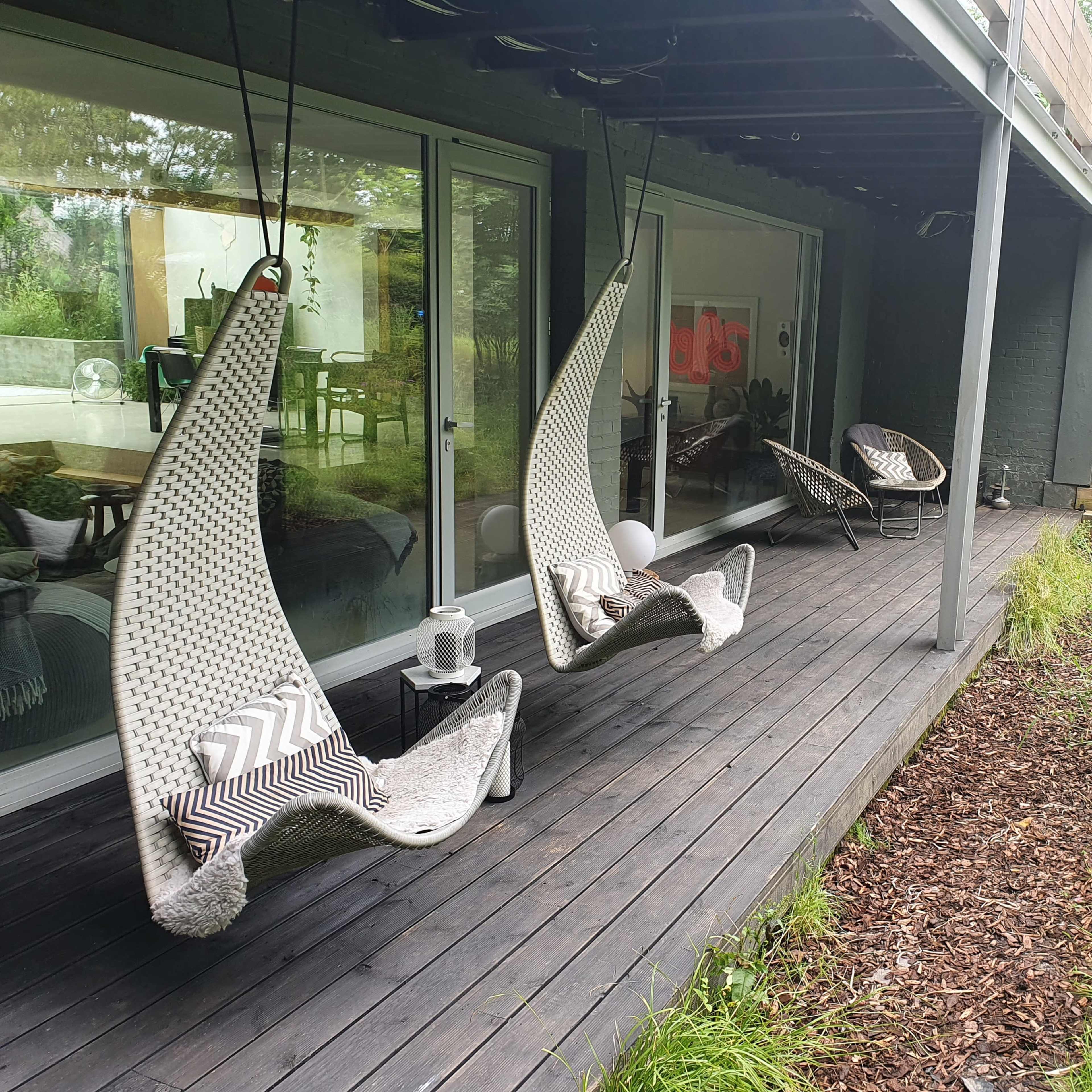 A wooden deck with two hanging chairs and a small table, surrounded by greenery and a view of a modern house.
