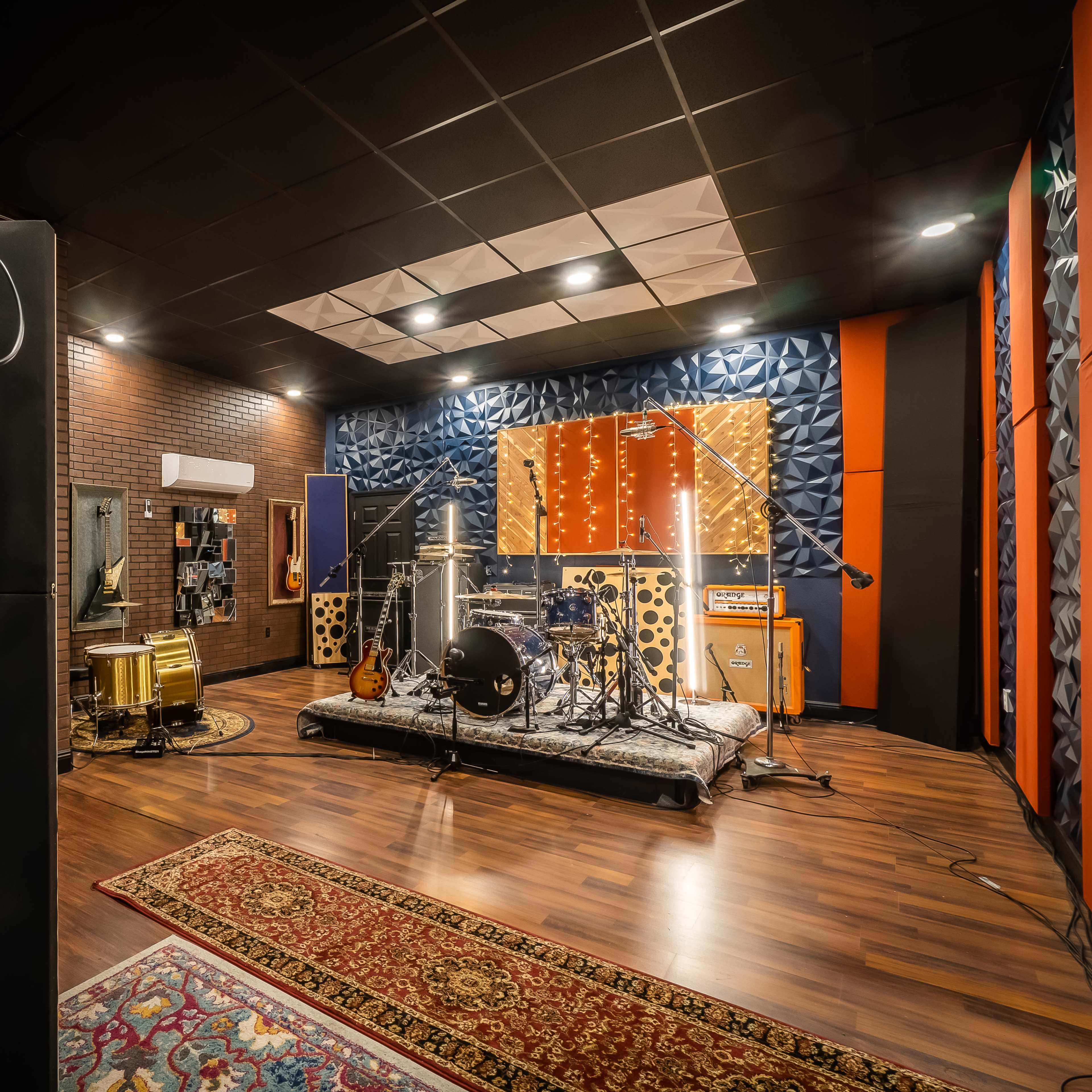 A music recording studio featuring microphones, drums, amplifiers, and soundproofing panels, along with a patterned rug on the wooden floor.