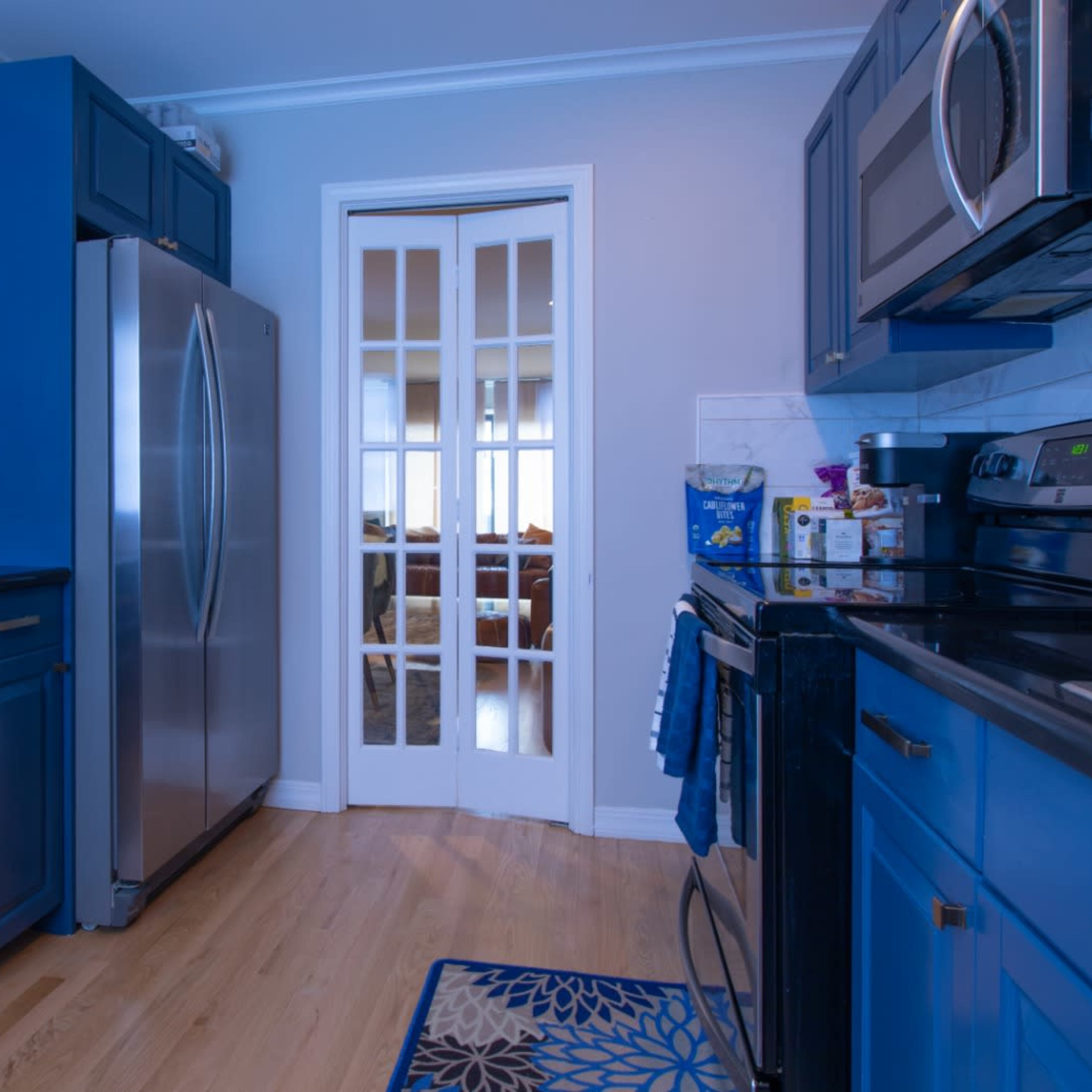 A modern kitchen features blue cabinetry, stainless steel appliances, and a view into a adjacent room through a set of glass doors.