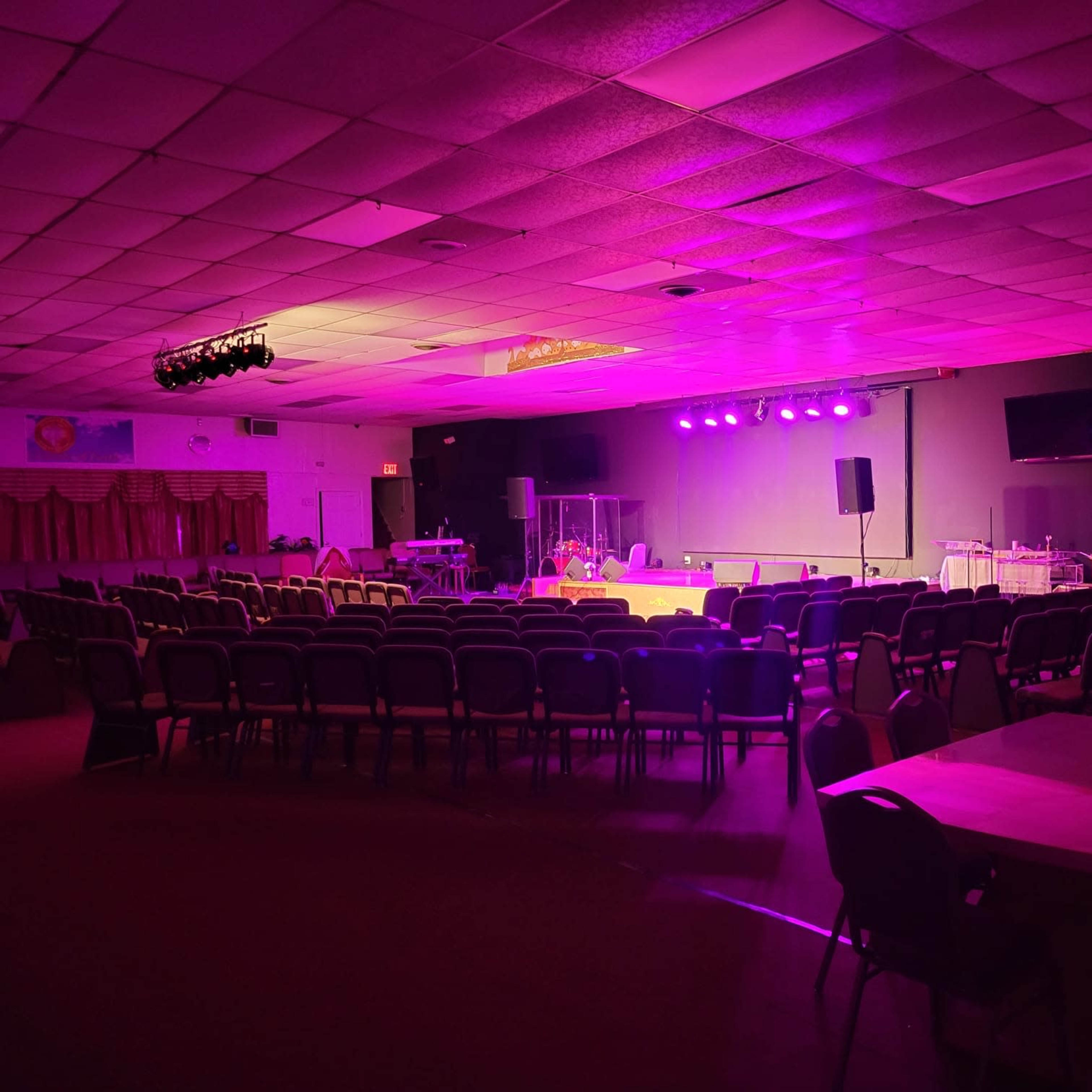 A dimly lit auditorium with rows of chairs facing a stage, illuminated by purple and white stage lights.