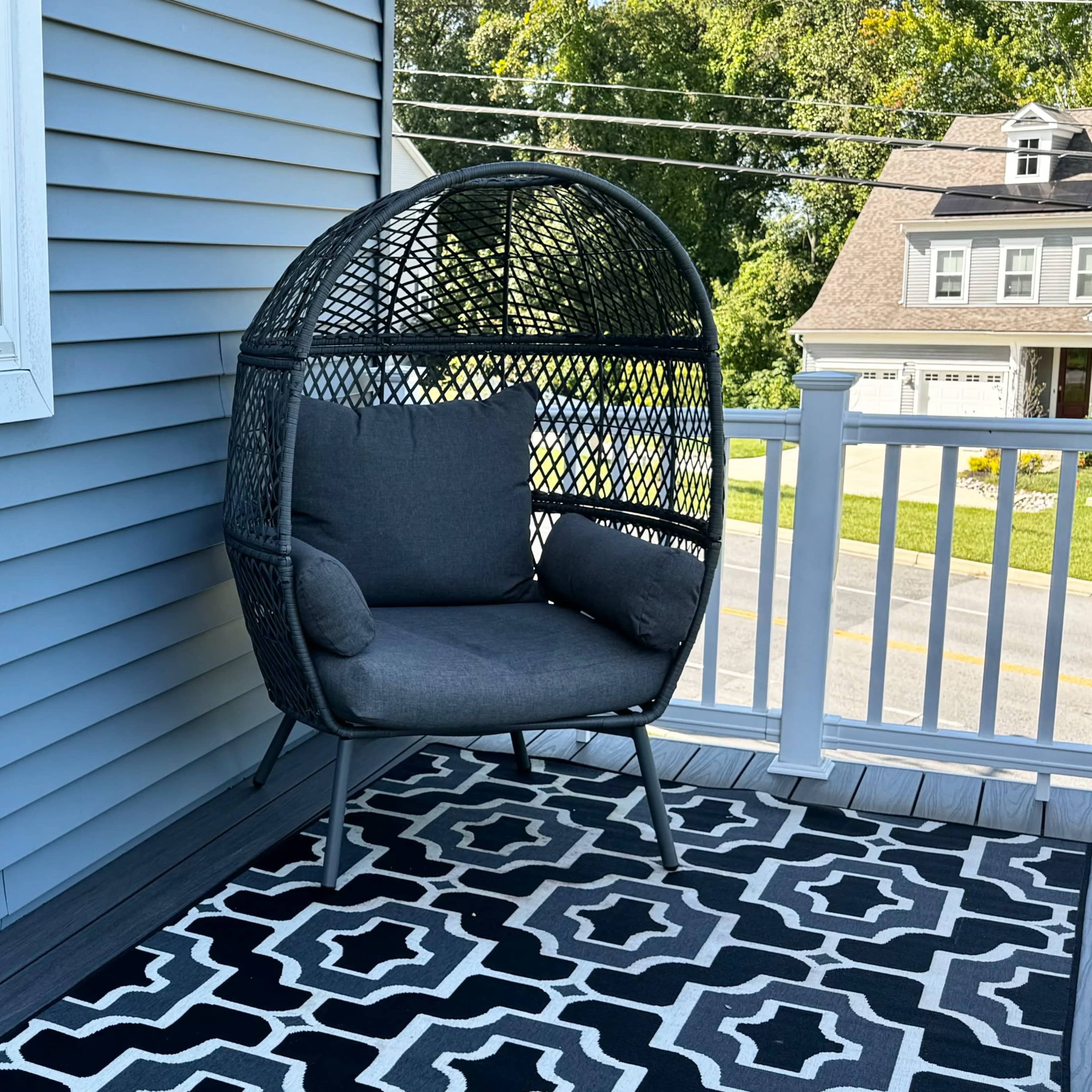 A gray, woven patio chair is positioned on a patterned outdoor rug next to a window on a house with blue siding.