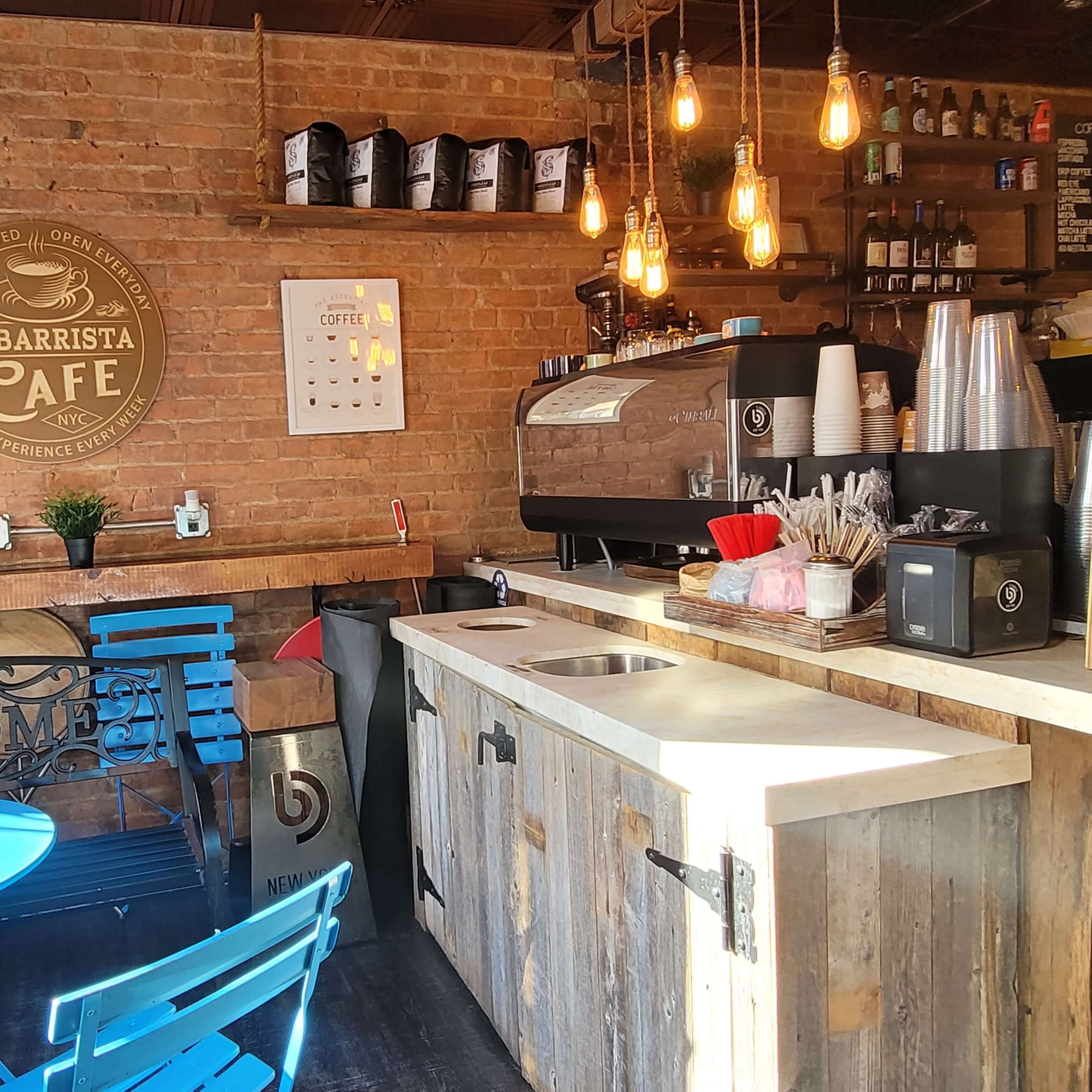 A cozy coffee shop features a brick wall, wooden counter, and blue tables with chairs near a coffee machine and menu board.