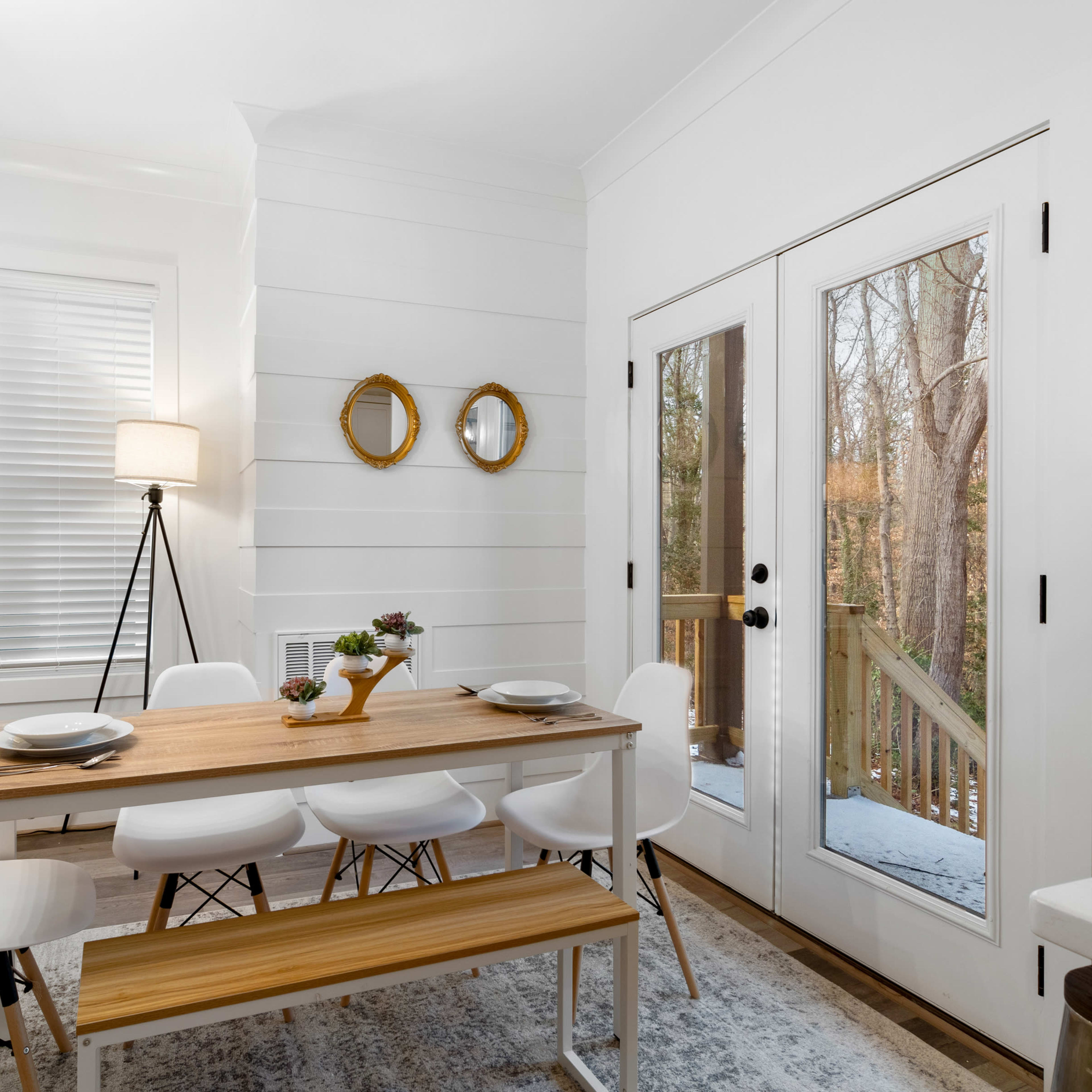 A bright dining area features a wooden table with white chairs, a floor lamp, and double glass doors leading to an outdoor deck.