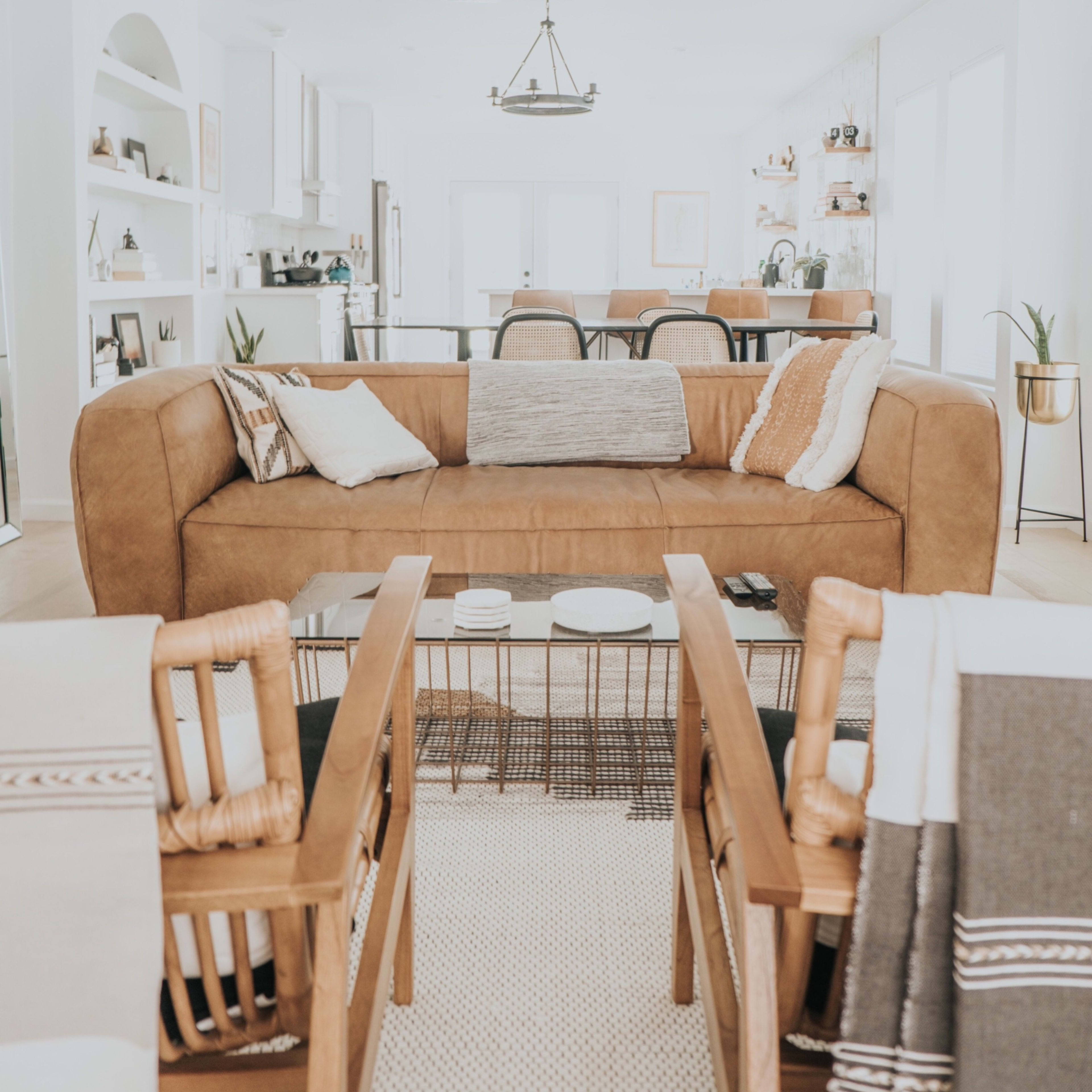 A modern living room features a large brown leather sofa, a coffee table with decorative items, and two wicker chairs, all arranged around a light-colored rug.